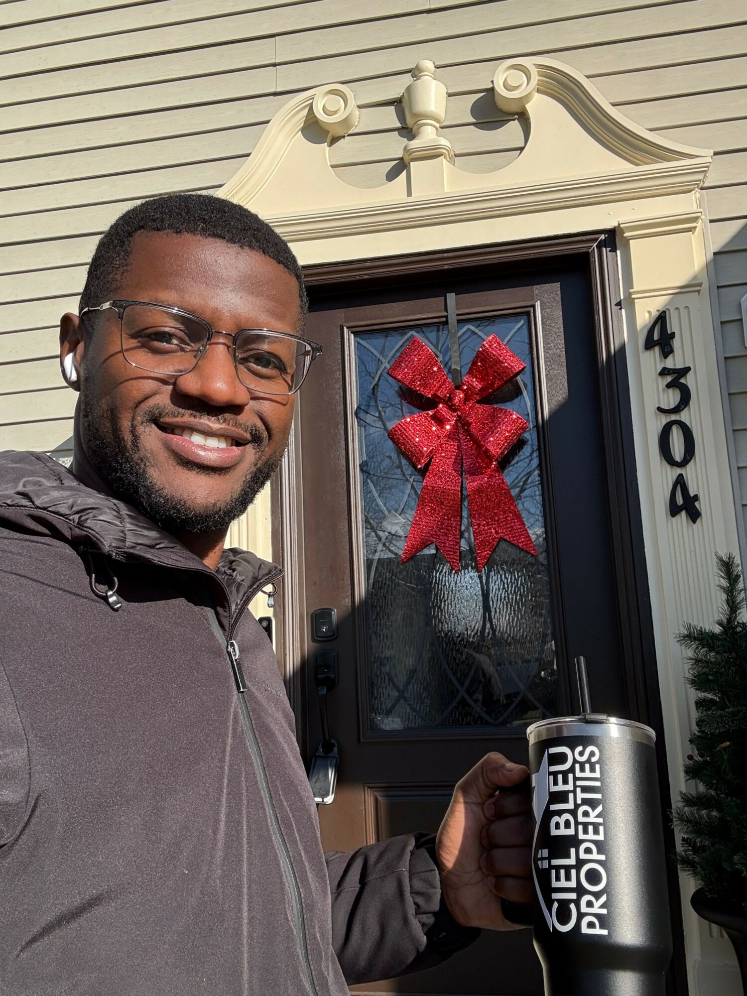 Man smiling outside house holding a can, decorated door with a red bow, house number 4304 visible.