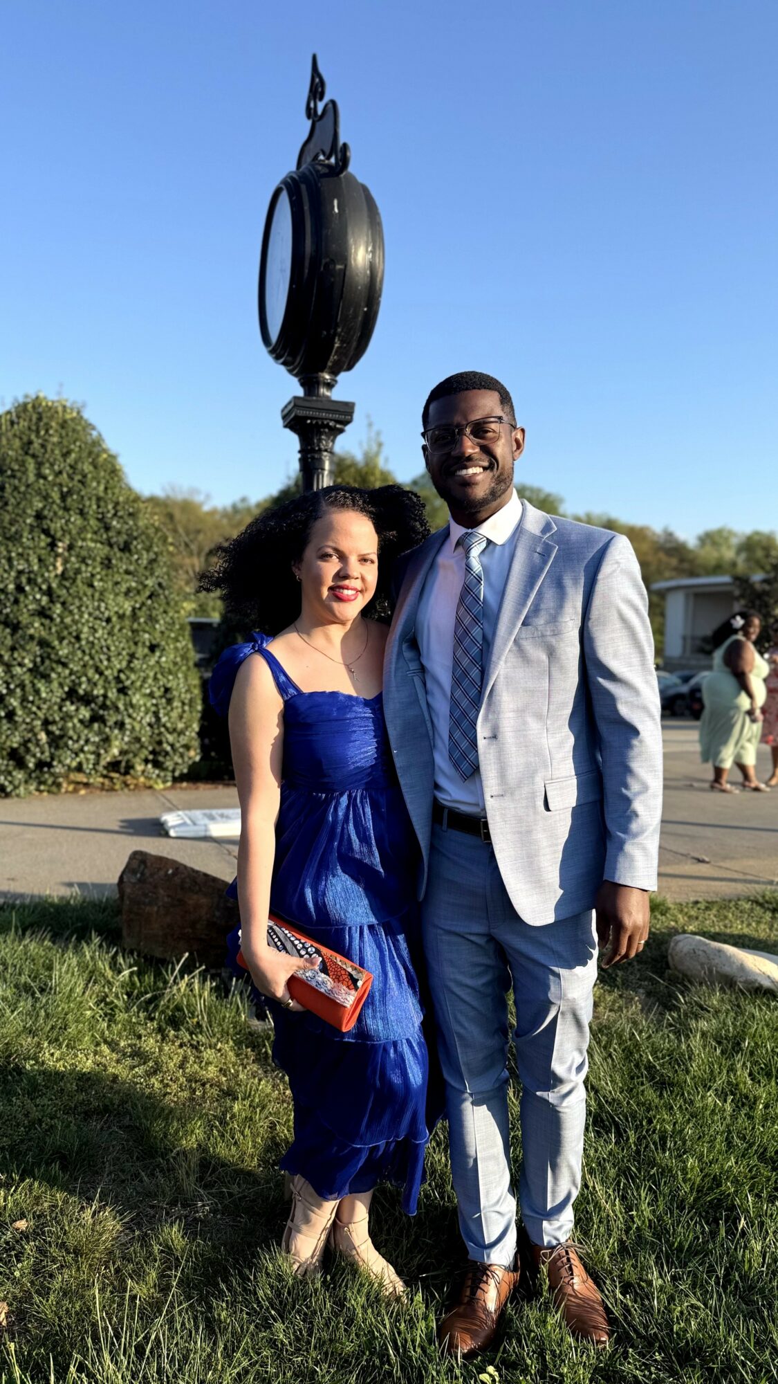 A smiling woman in a blue dress and a man in a light gray suit stand outdoors near a sculpture, smiling at the camera.