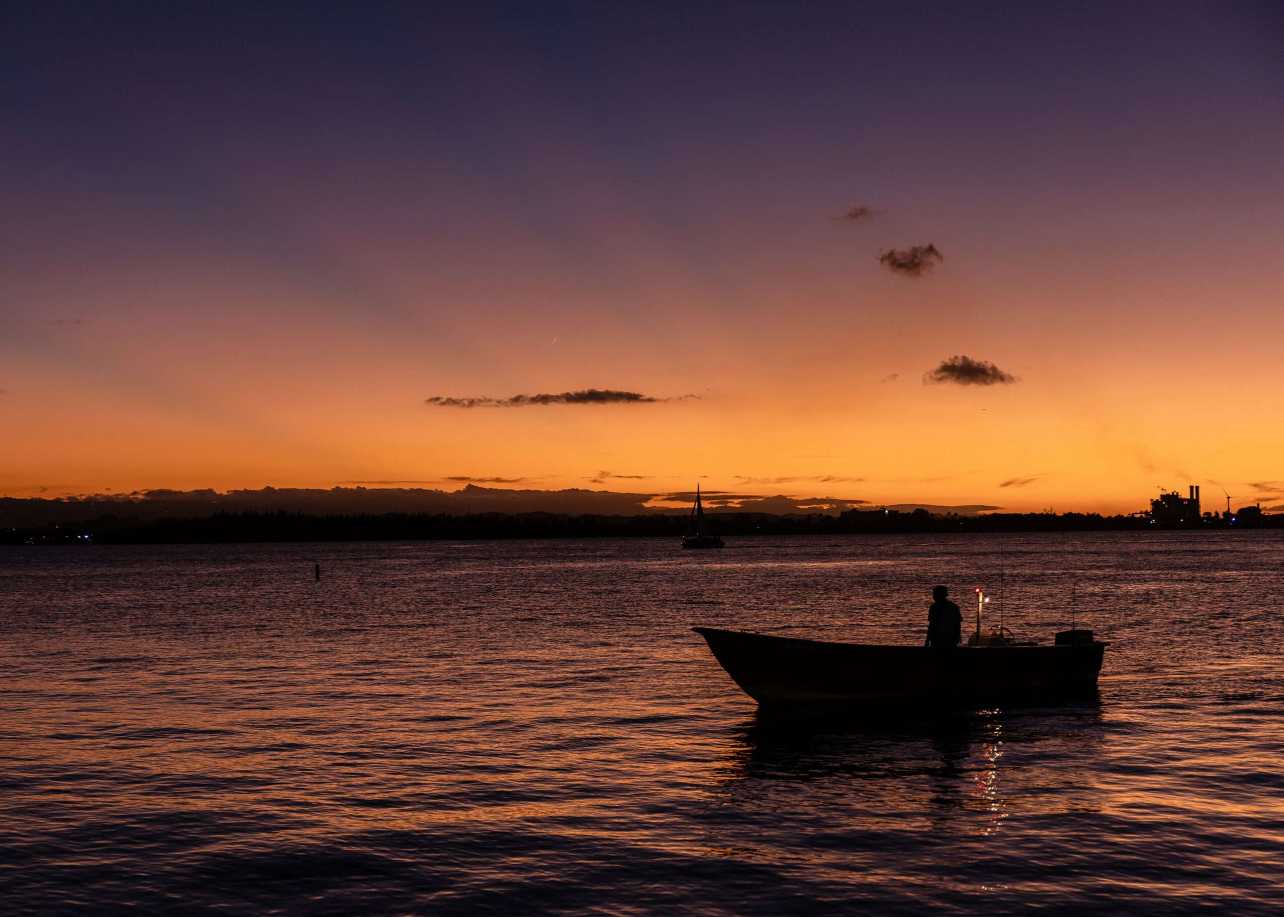 Silhouette of a boat with a person on water during sunset with colorful sky and distant horizon.