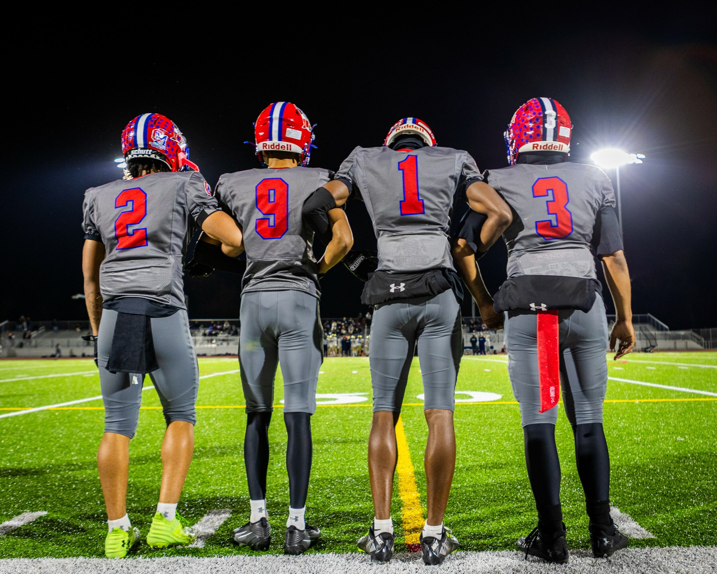 Four football players stand on a field at night, facing away, wearing gray uniforms with red and blue helmets.