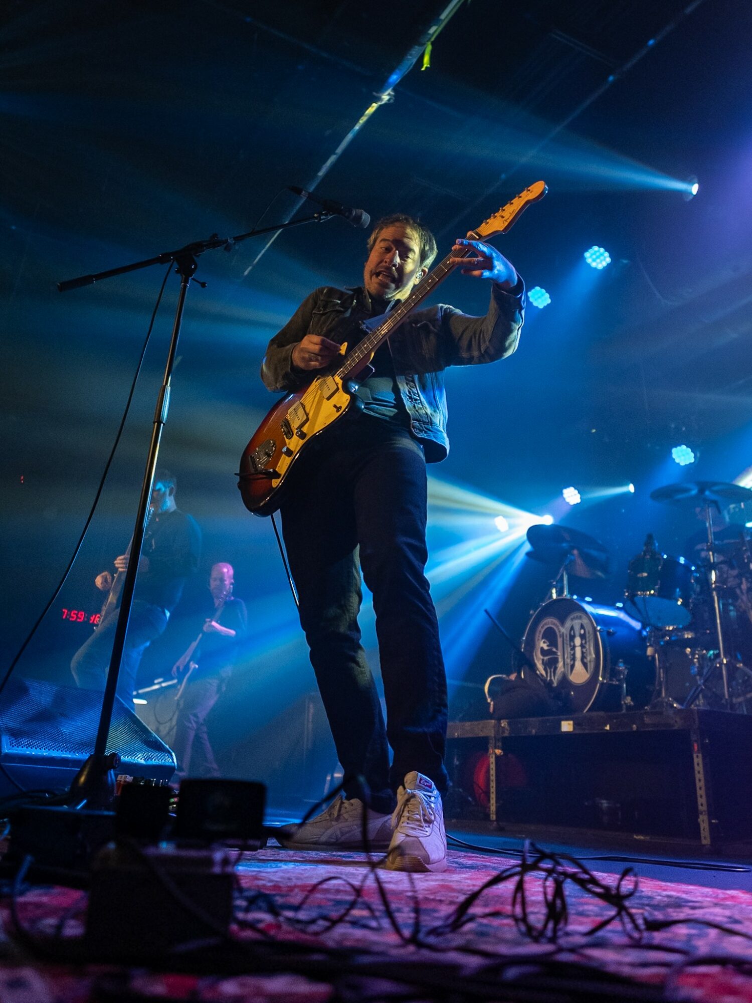 Female guitarist performing on stage with blue and white lighting, microphone stand, and band members in background.