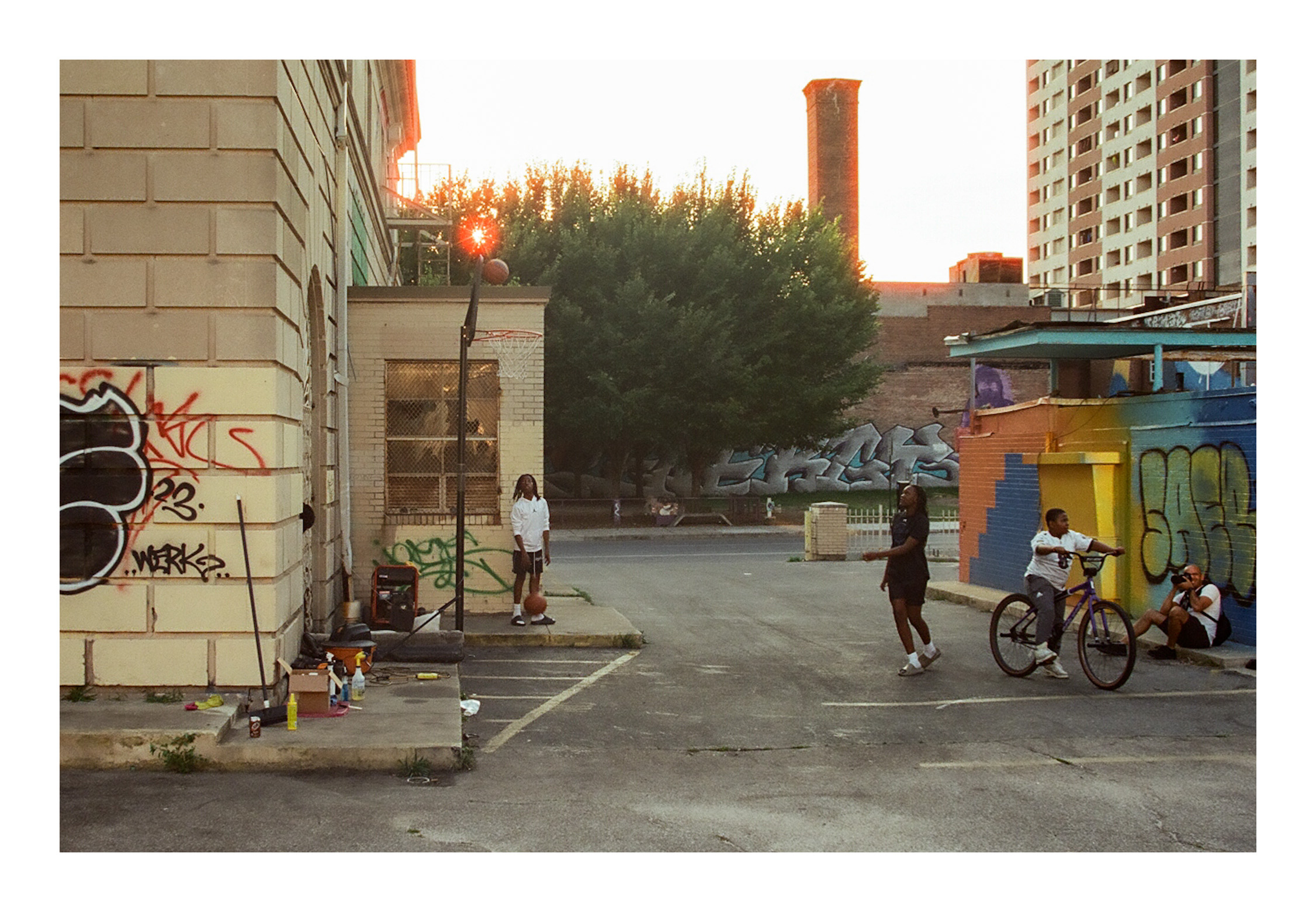 Urban scene with people, bicycles, graffiti, and buildings, including a tree and a colorful wall, during sunset.