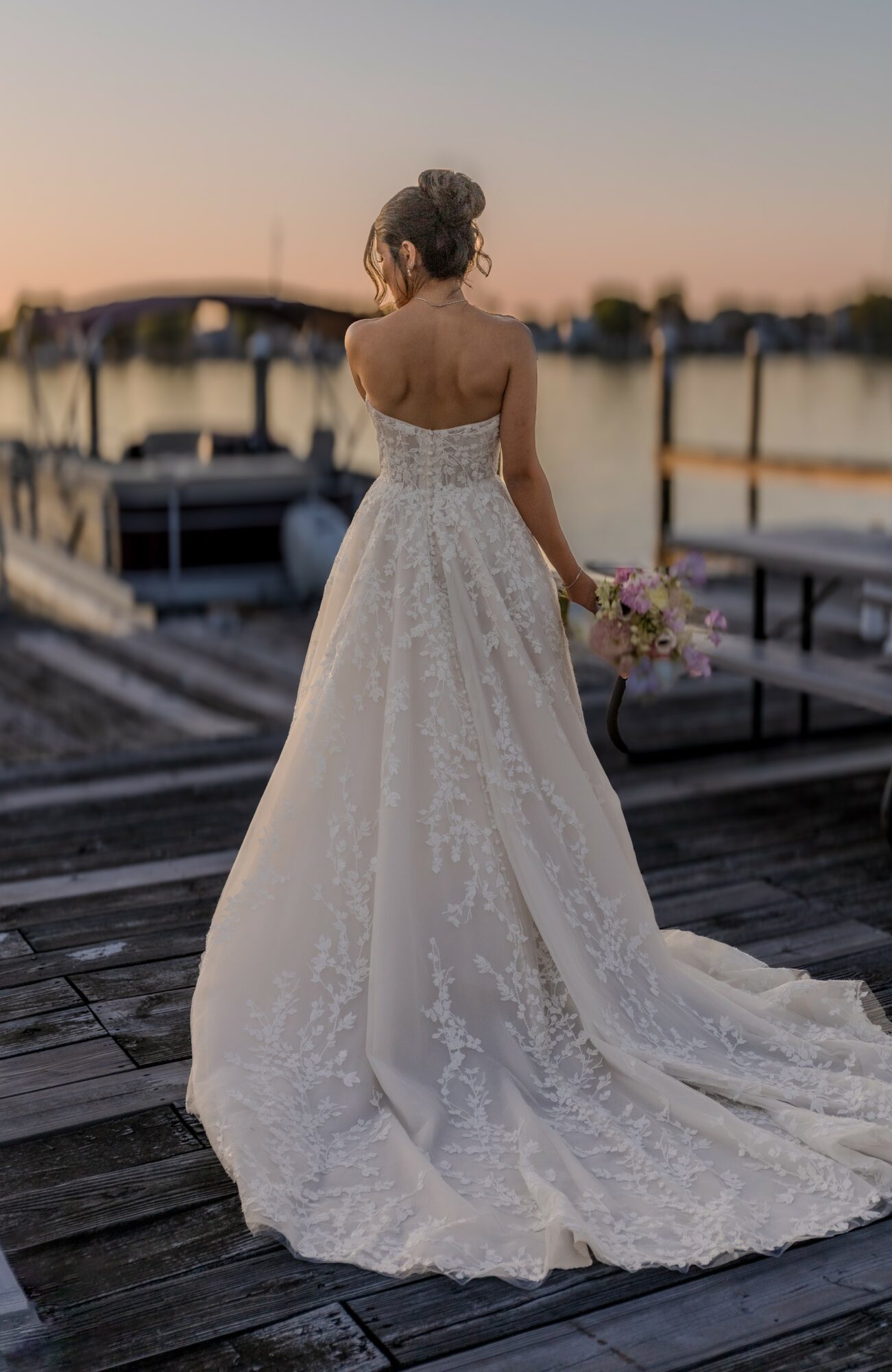 Woman in wedding dress holding bouquet, standing on dock near boats at sunset, back view.