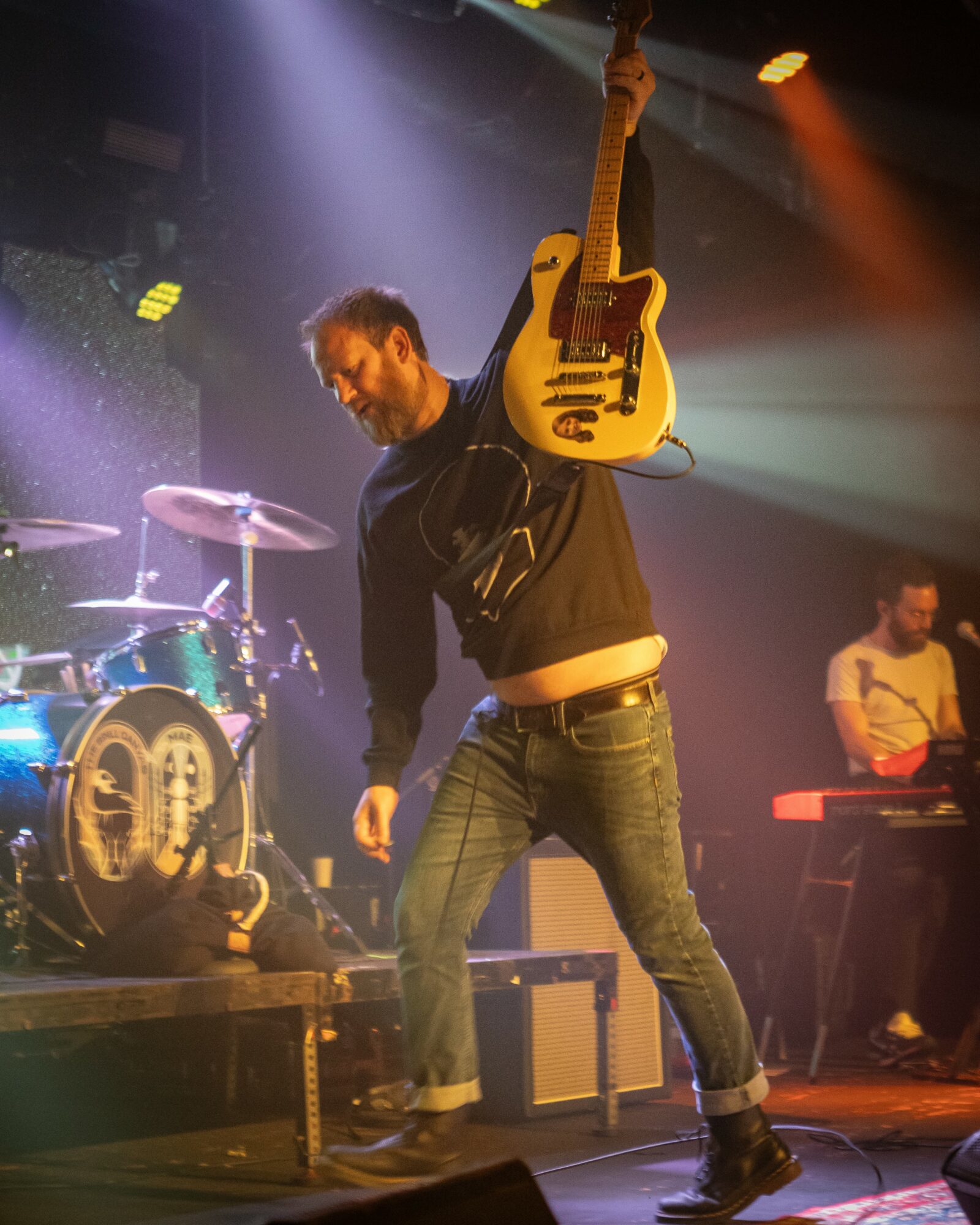 Musician on stage holding a yellow electric guitar over shoulder, with a drum set and another musician in background.