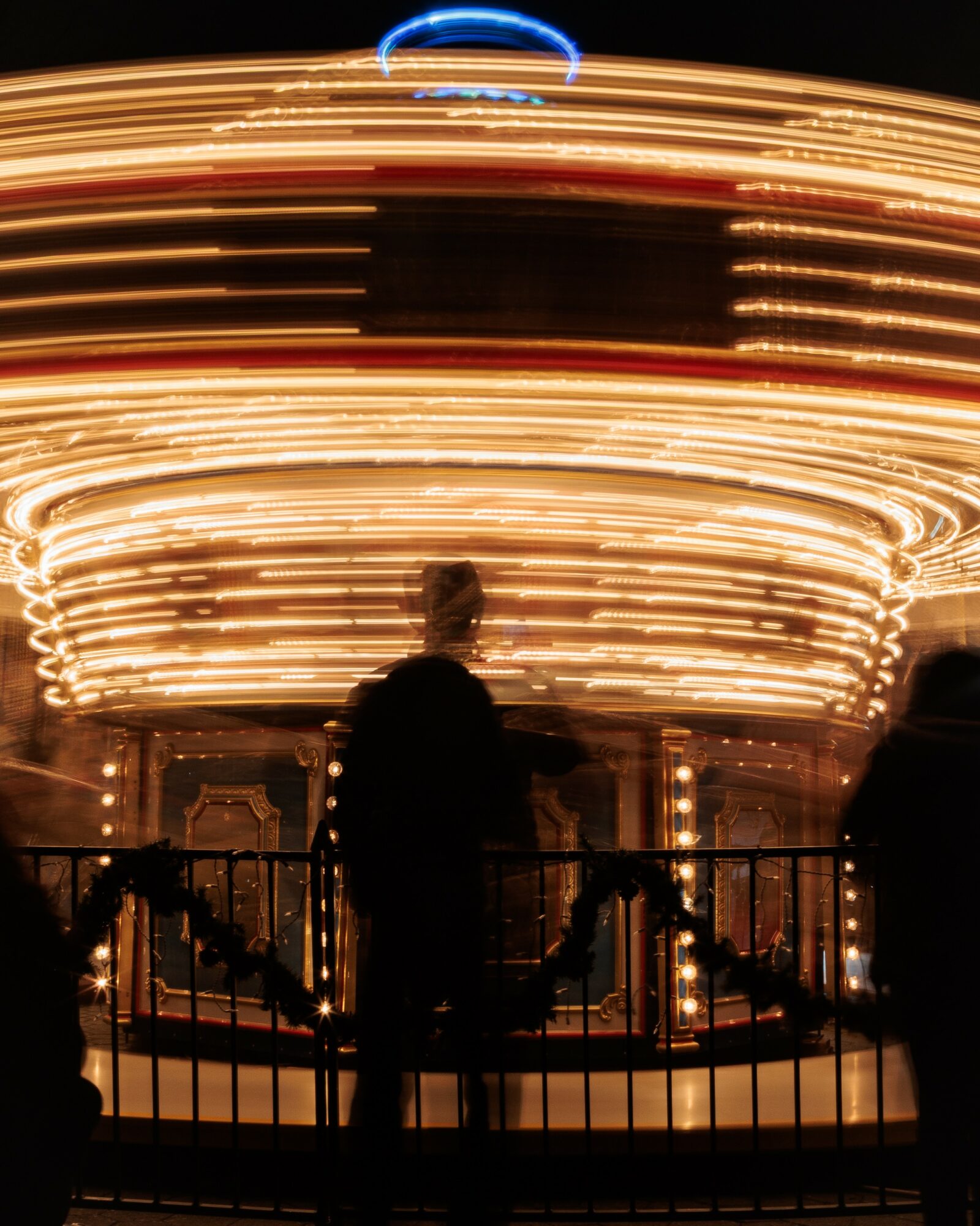 Silhouettes of people in front of a brightly lit carousel at night, with light trails creating a circular pattern.