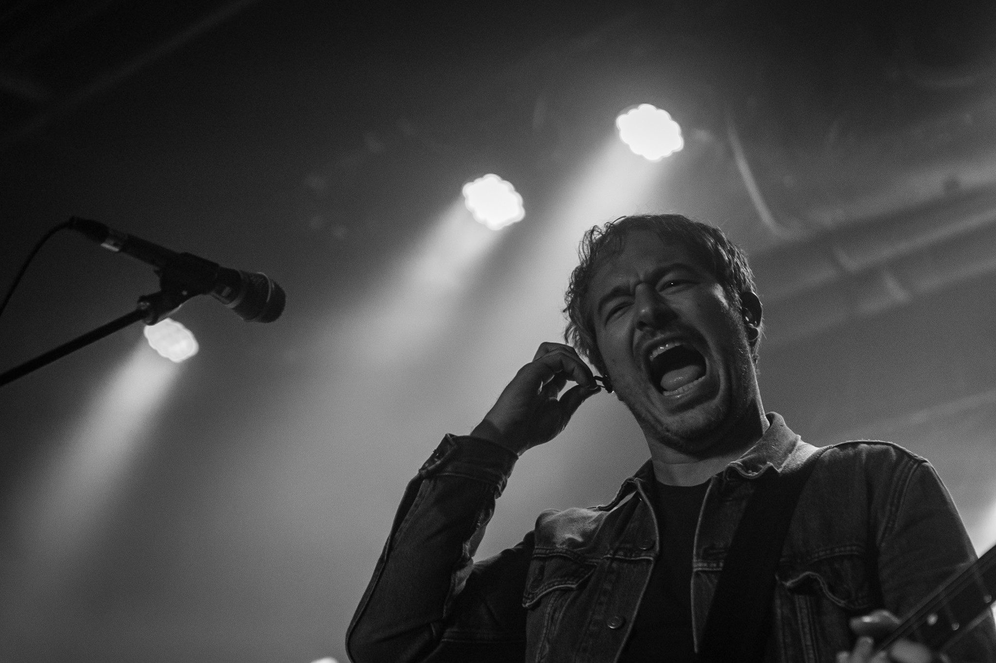 Person singing passionately on stage with microphone, stage lights overhead, black and white image.