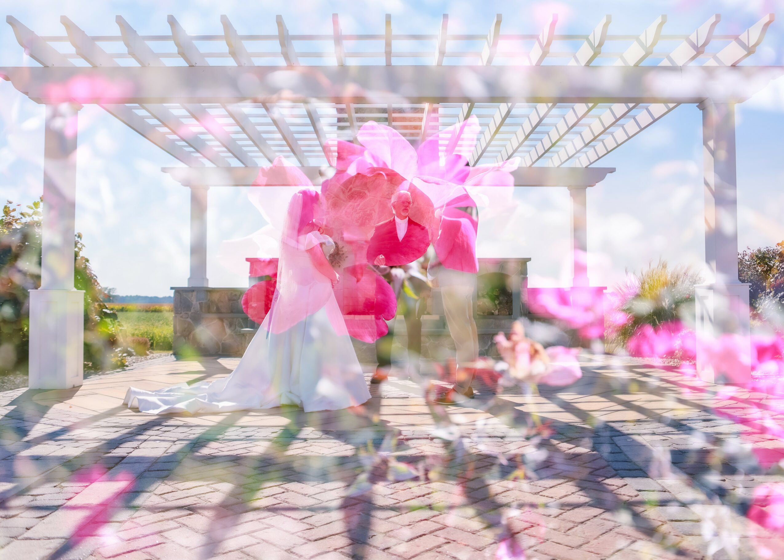 Bride in wedding dress under a pink floral arch on a patio with a pergola and sky background.