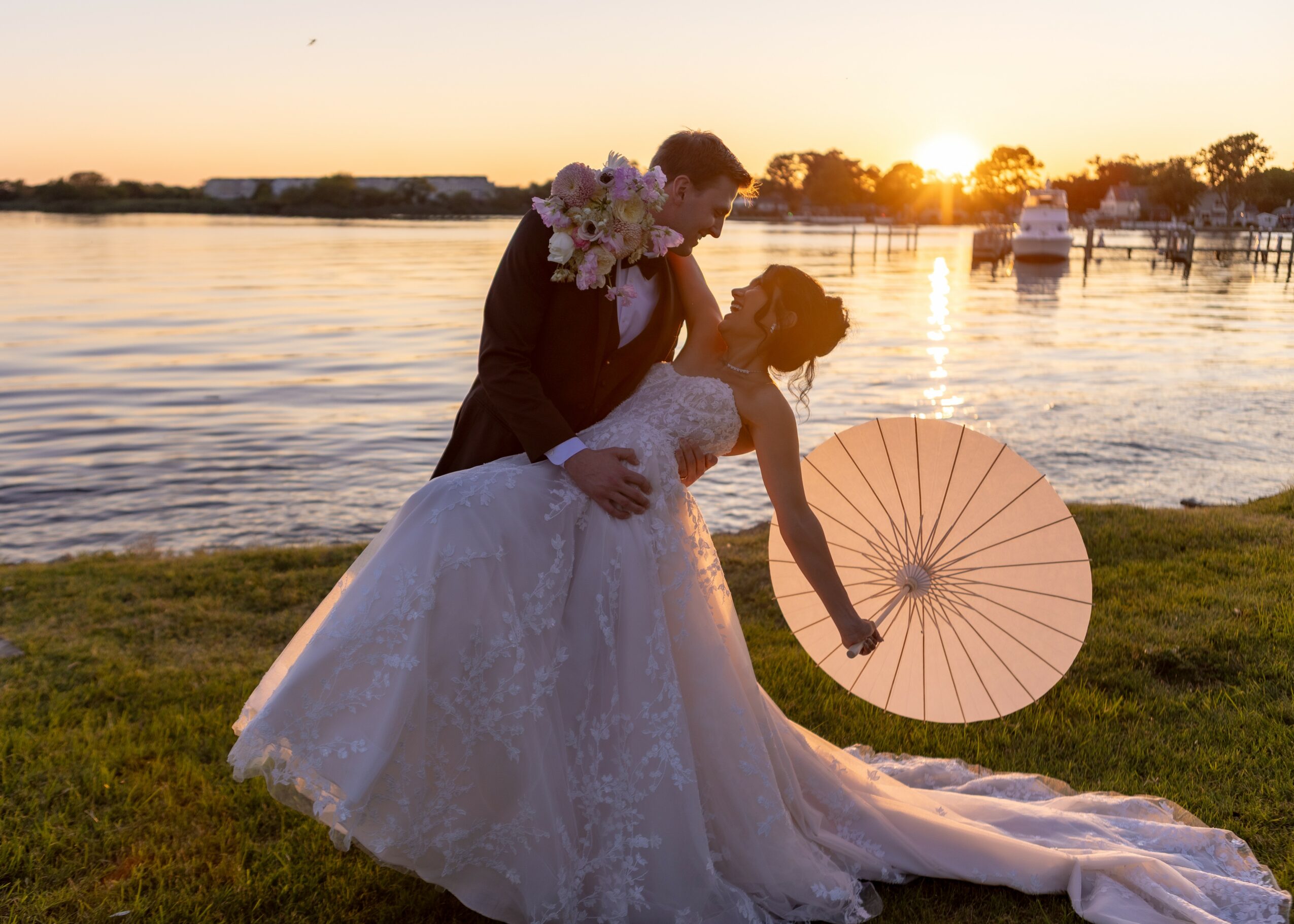 Couple in wedding attire by water, holding a pink parasol, sunset background, boats on water, grassy area.