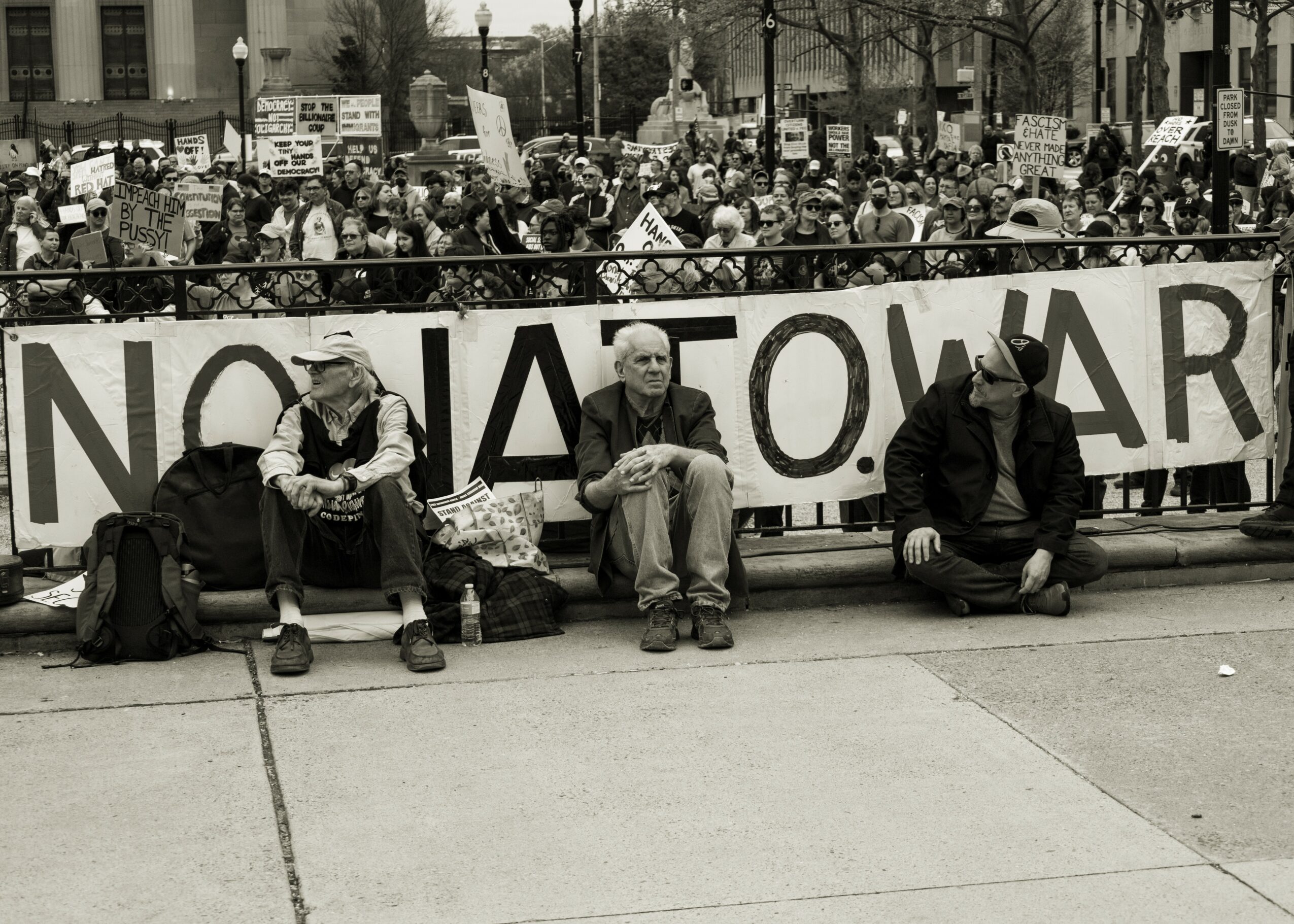Four people sitting on the ground in front of a large protest banner, with a crowd holding signs behind them.