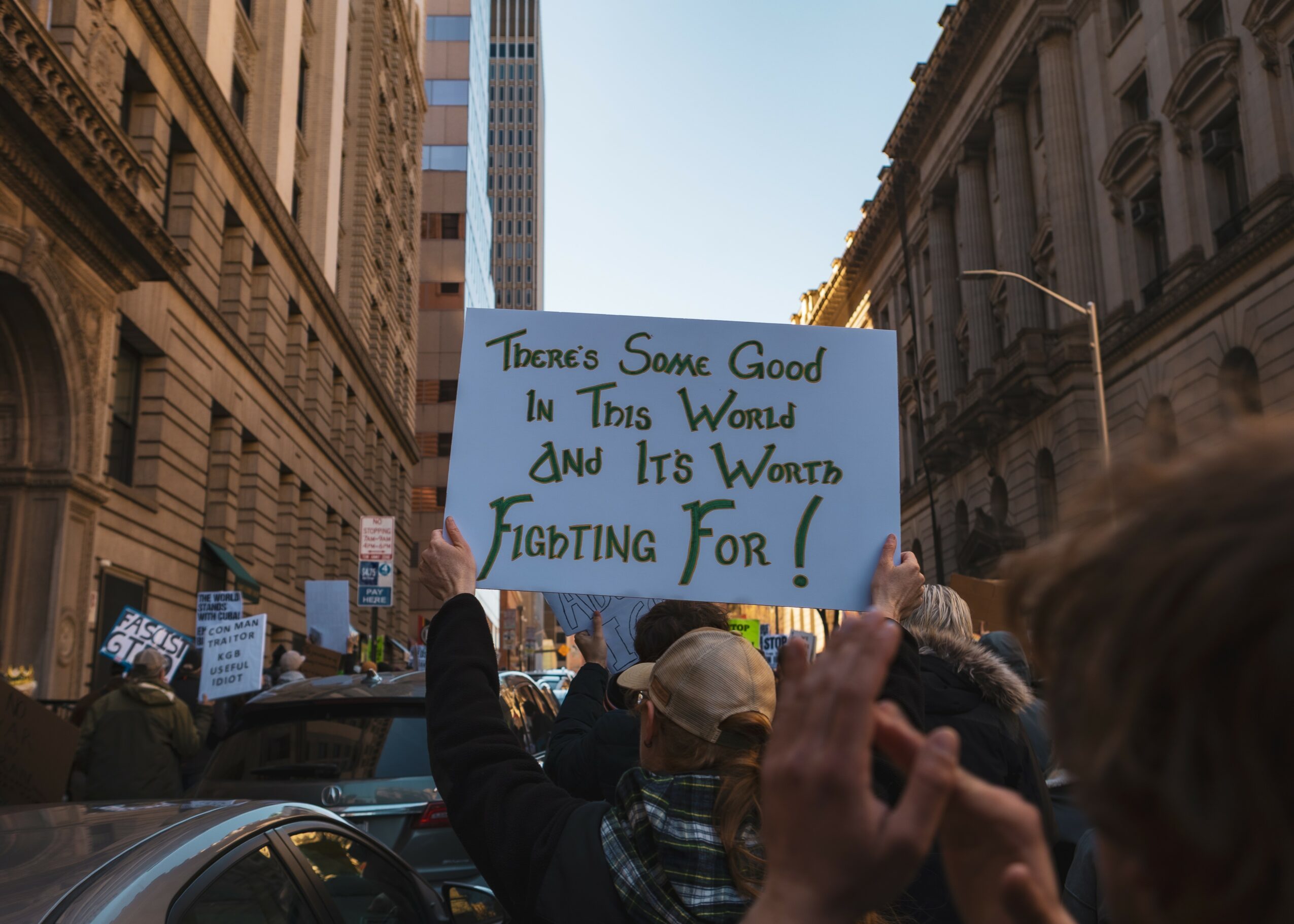 Crowd of people protesting on city street, holding signs, with tall buildings on both sides.