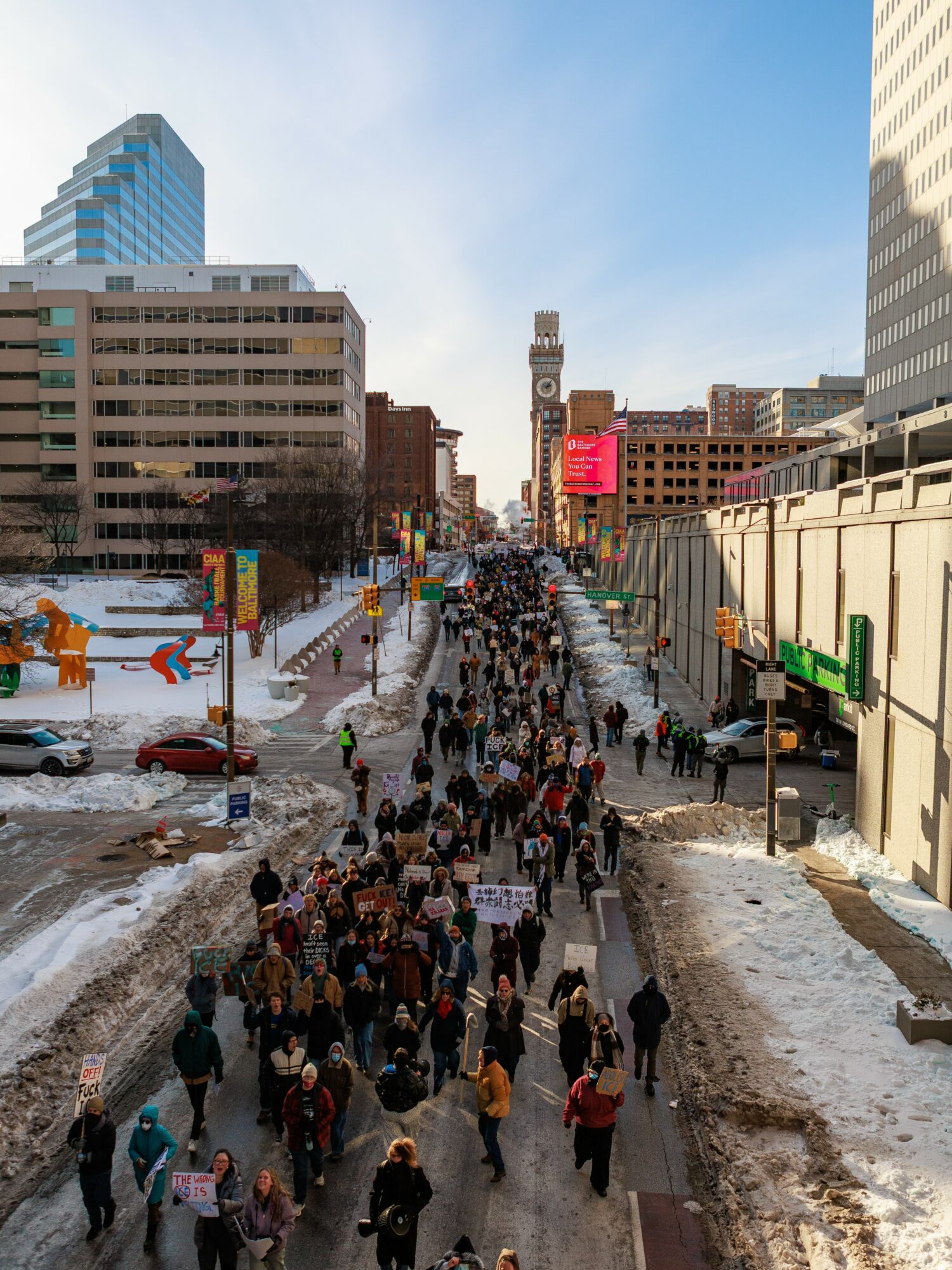Crowd walking on a city street with tall buildings, some snow on the ground, and a clear sky.