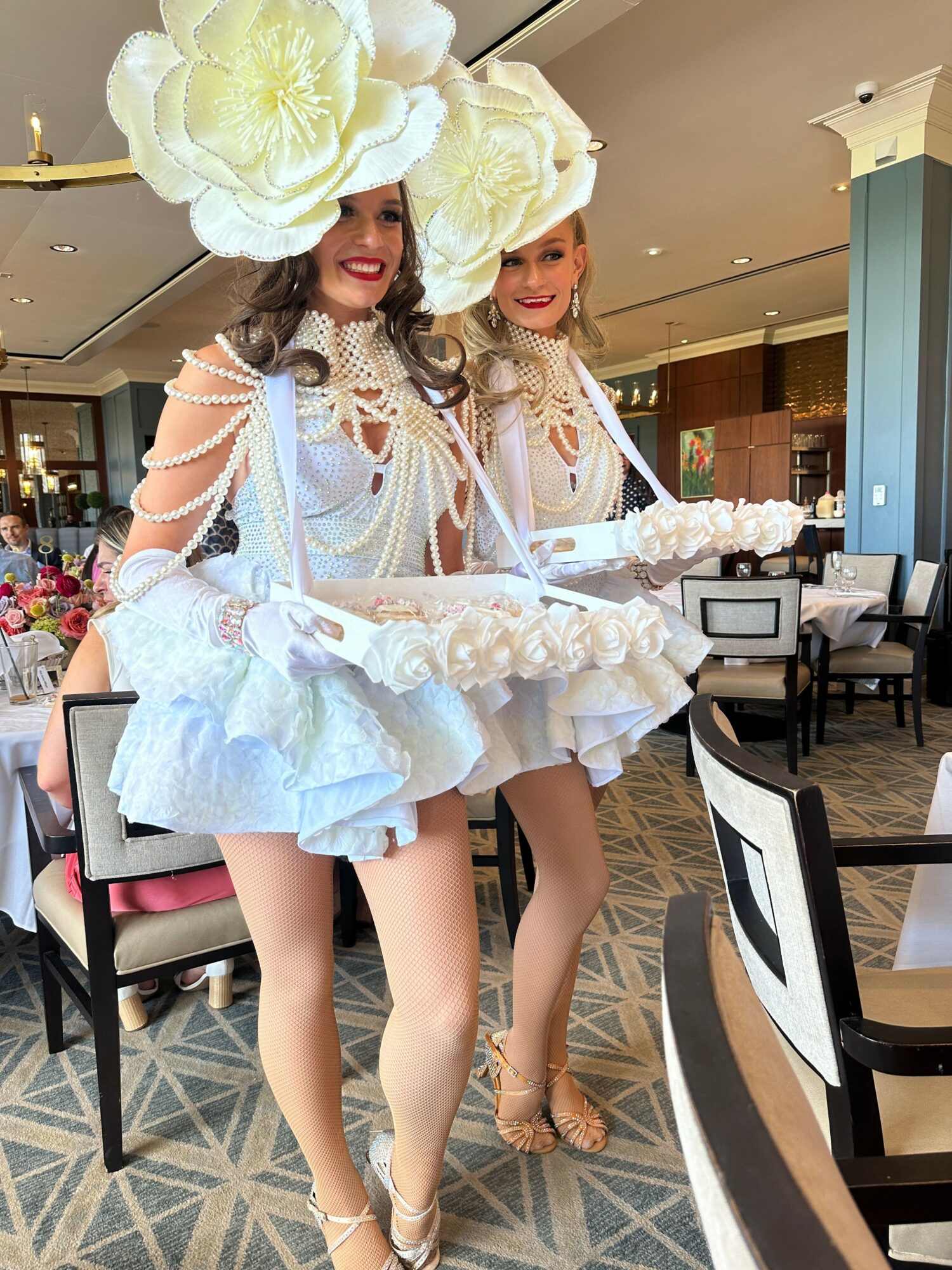 Two women in white dresses with large floral headpieces and gloves, standing in a restaurant or cafe setting.