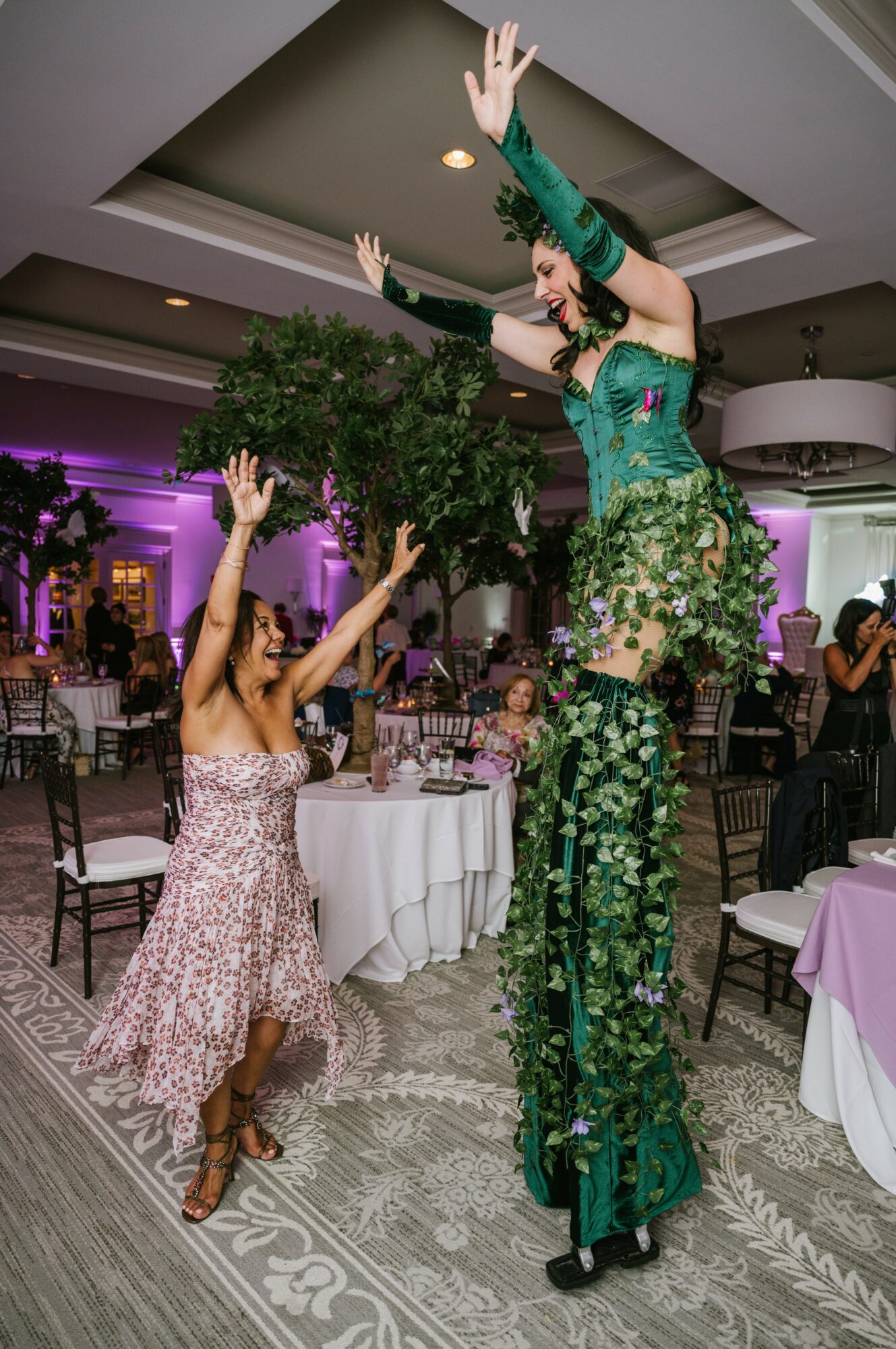 Two women dance indoors, one on stilts wearing a green floral outfit, the other in a pink patterned dress, with arms raised.