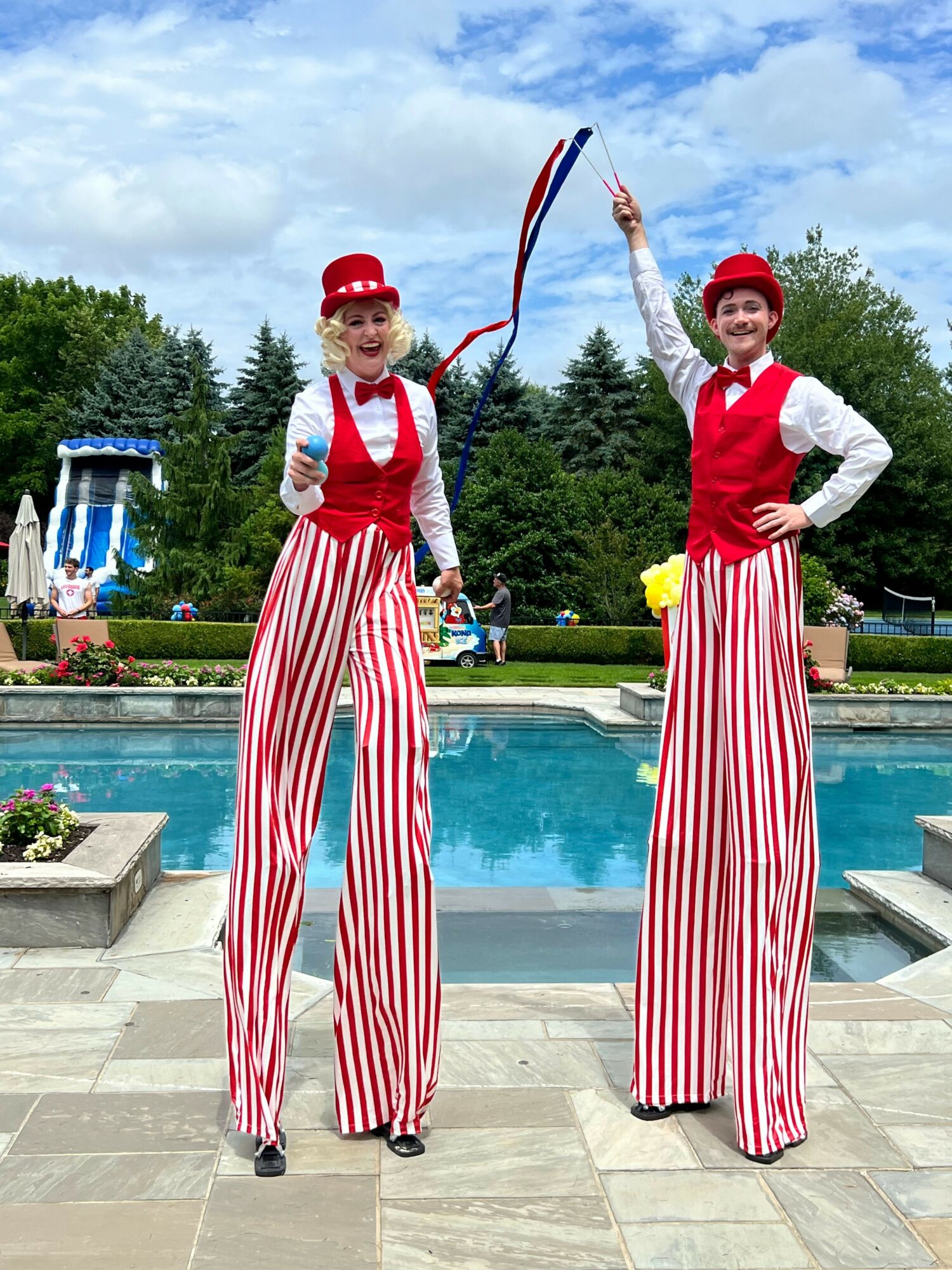 Two performers in red and white striped pants, red vests, white shirts, red hats, standing by a pool outdoors.