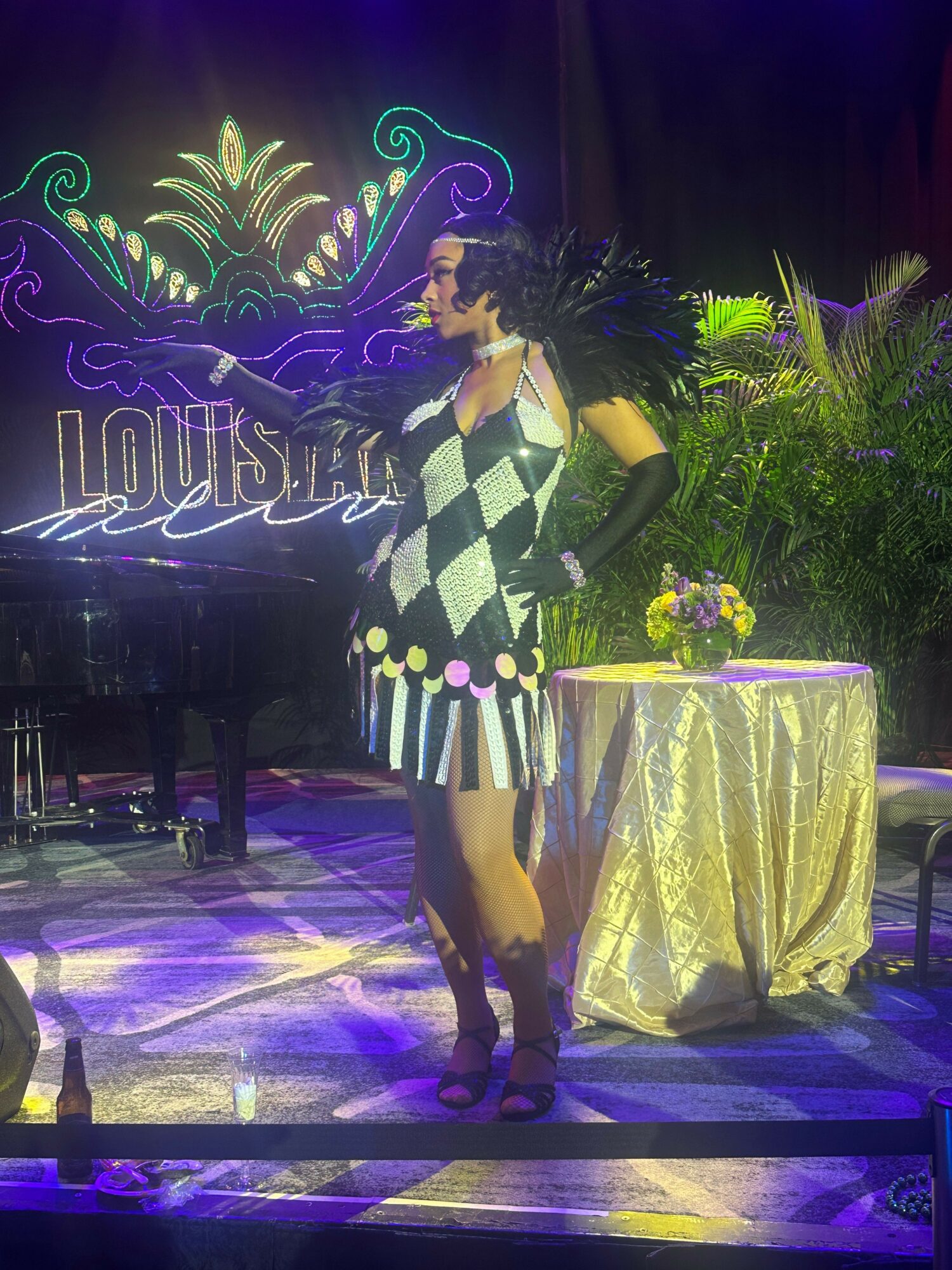 Person standing on stage with decorative backdrop, wearing a checkered dress and gloves, next to a table with a yellow cloth and flowers.