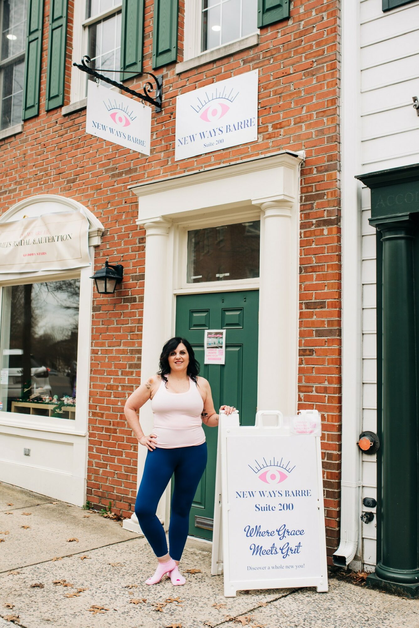 Woman standing outside a brick building with a green door and signs, smiling, holding a paper, sunny day.