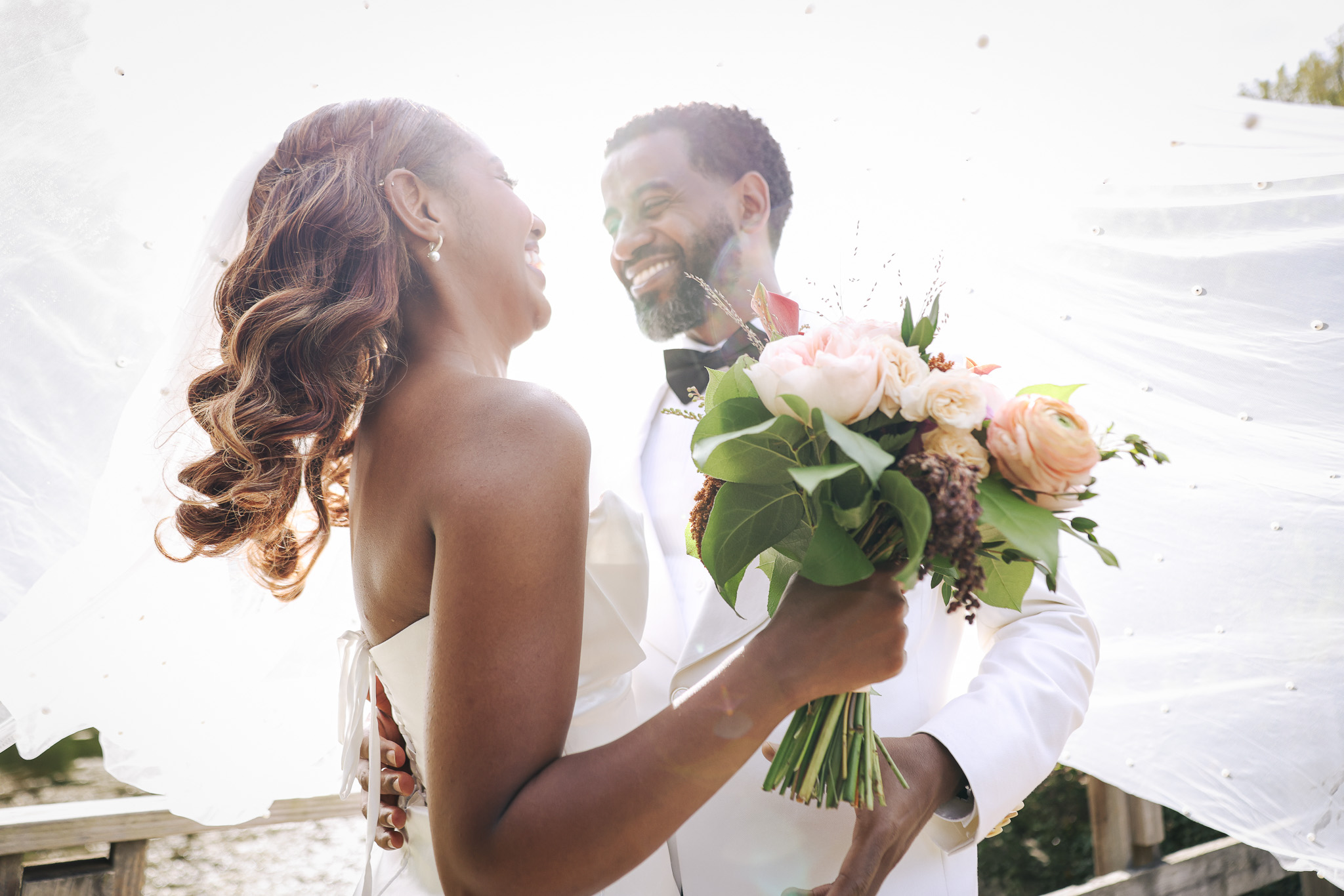 Bride and groom smiling at each other, bride holding a bouquet of pink and white flowers, outdoors setting.
