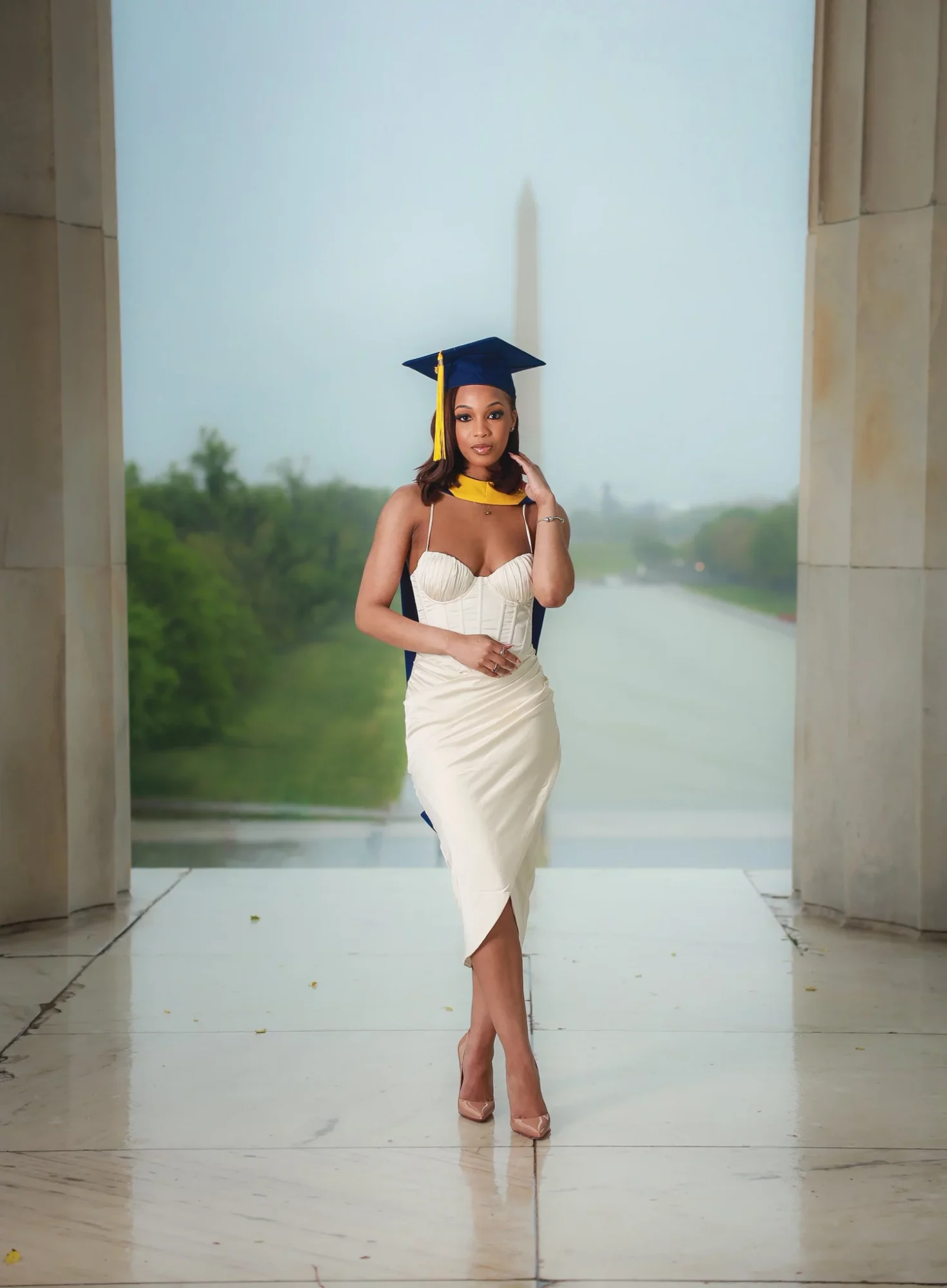 Woman in graduation cap and gown standing in front of large window with greenery outside.