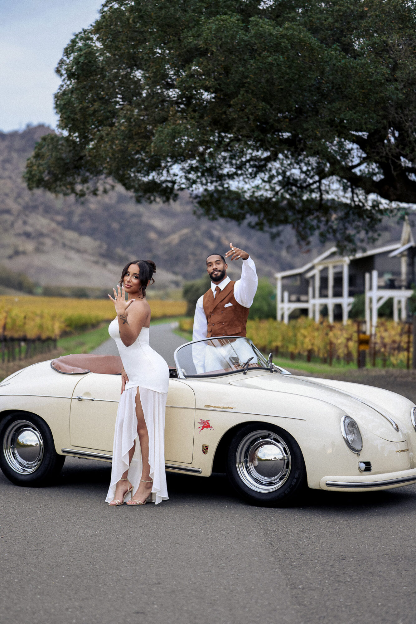 A woman in a white dress and a man in a vest and shirt stand beside a vintage cream-colored convertible car outdoors with trees and a building in the background.