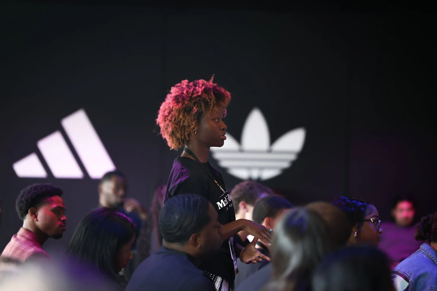 Young person with curly hair speaking to a group in a dark room with Adidas logos in background.