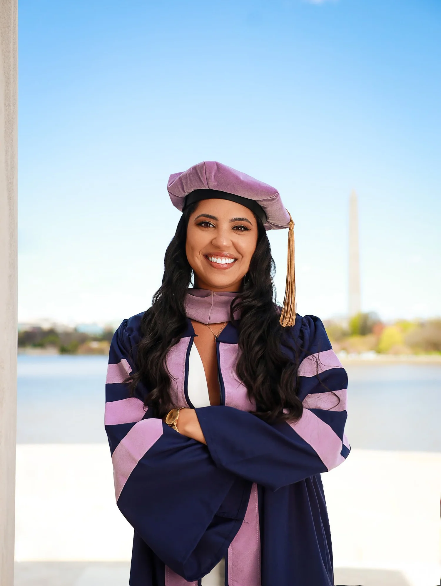 Woman in graduation gown and cap smiling outdoors with water and trees in background.
