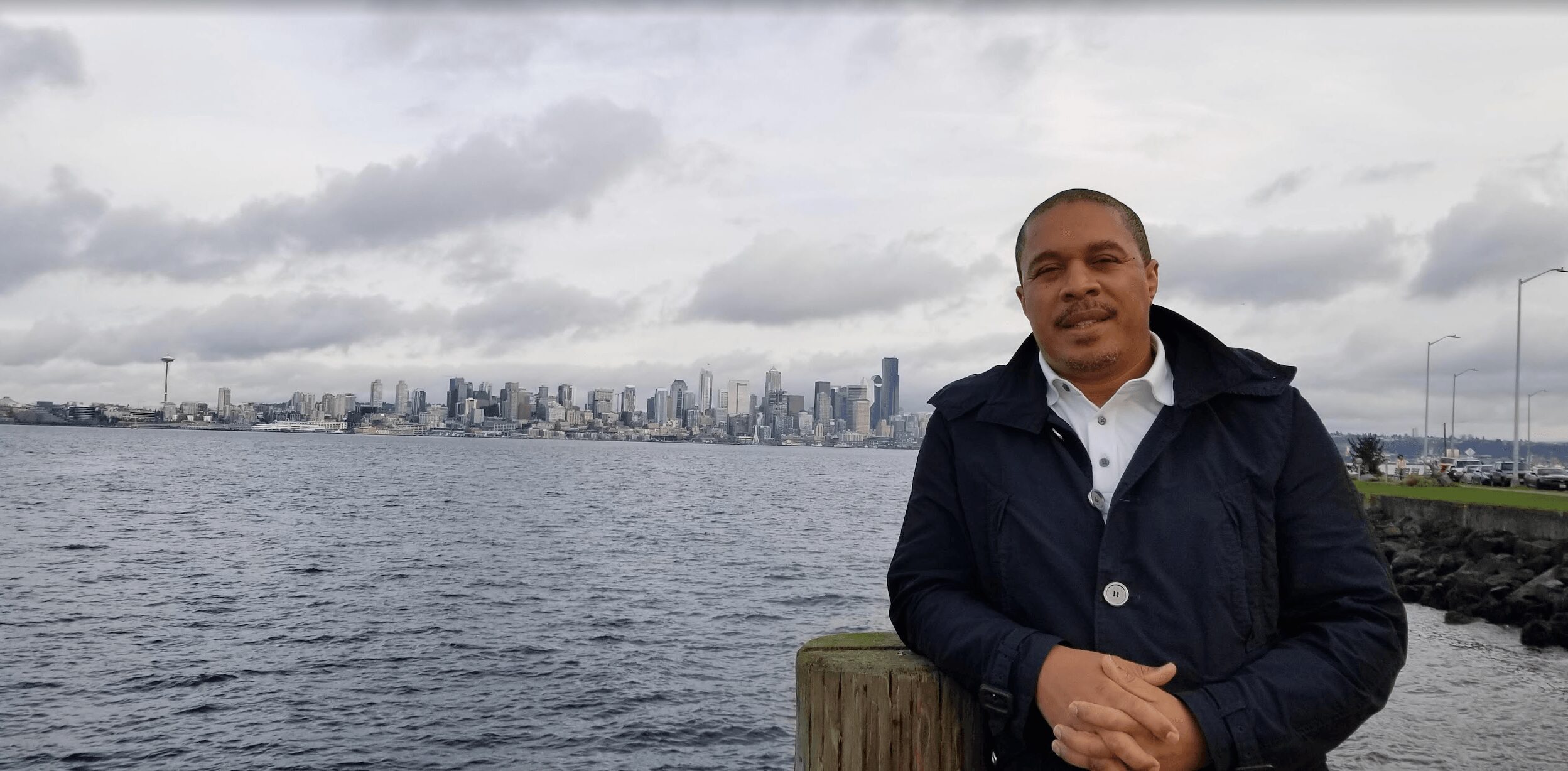 Man standing near water with city skyline in the background on cloudy day.