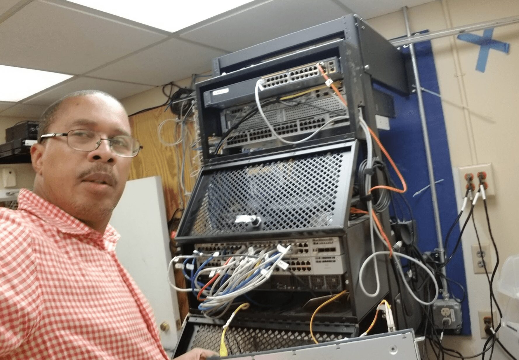 Man with glasses and red checkered shirt standing next to a server rack with cables and electronic equipment.