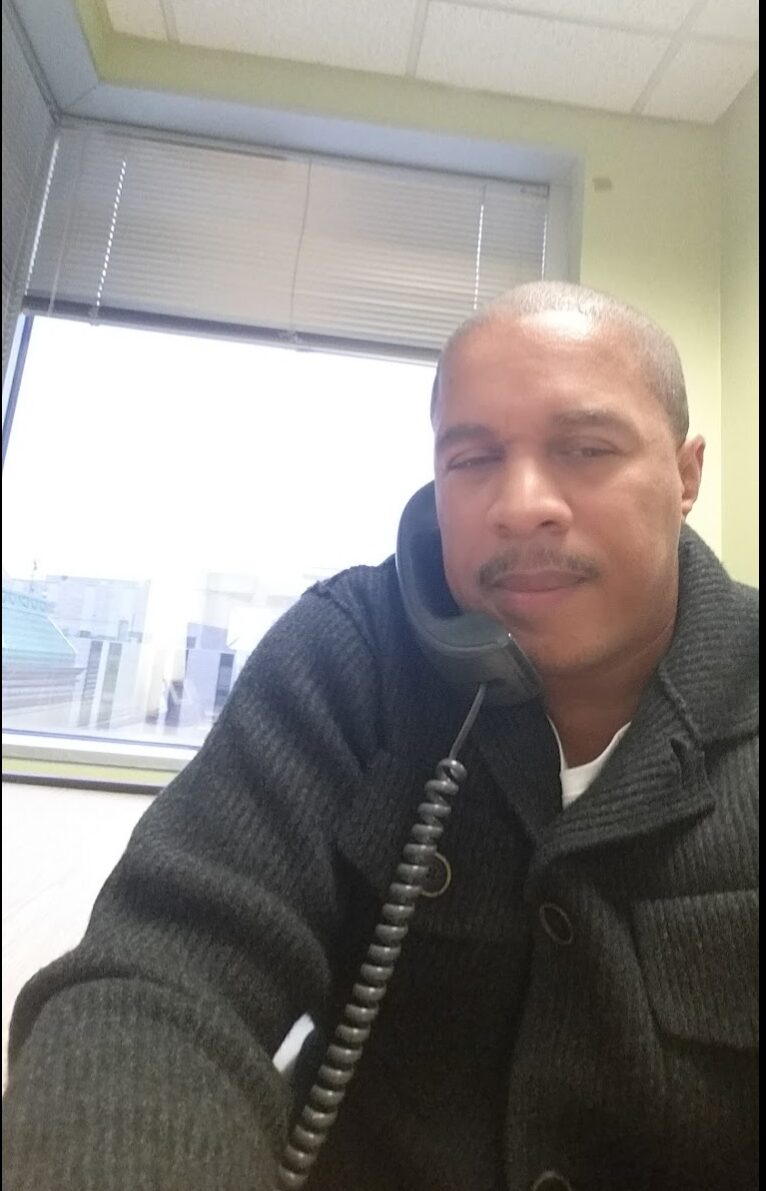 Man with short hair and mustache wearing a dark sweater, sitting indoors near a window with blinds, cityscape outside.