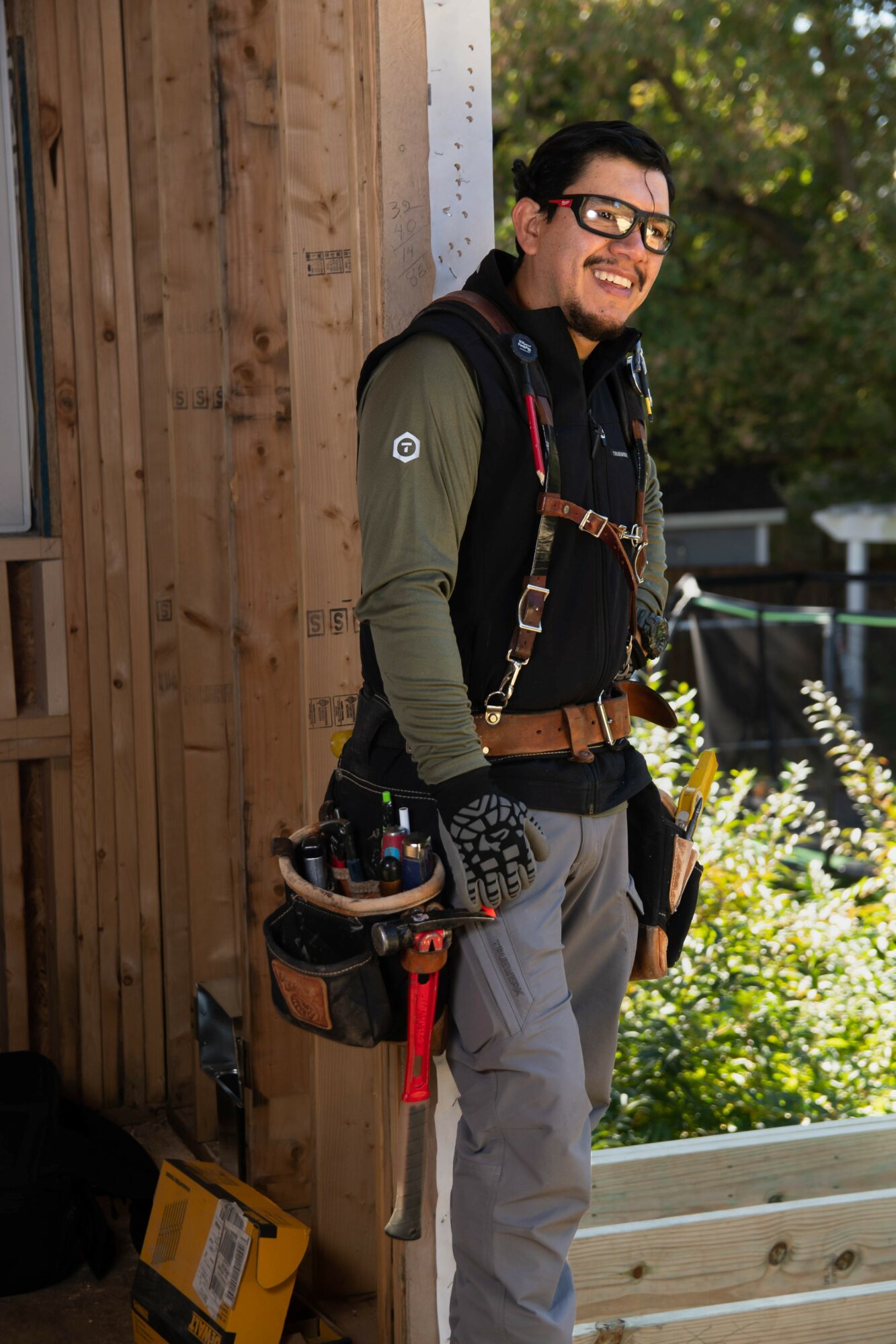 Man with glasses and a beard smiling outdoors, wearing a green jacket and carrying tools, standing near a wooden structure.