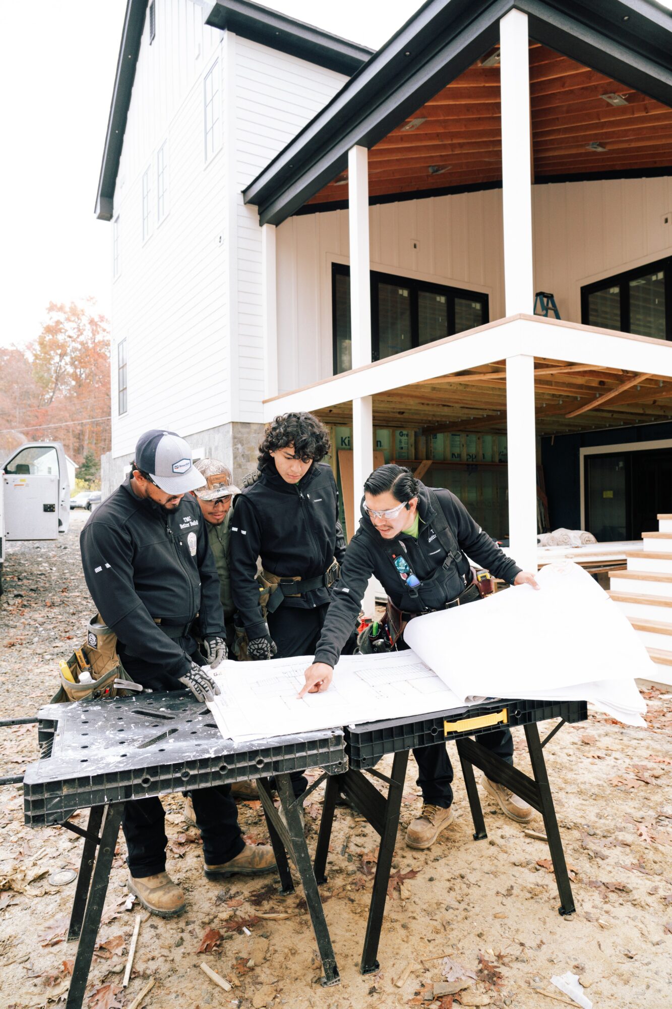 Four people examine blueprints on a black table outdoors near a modern house under construction.