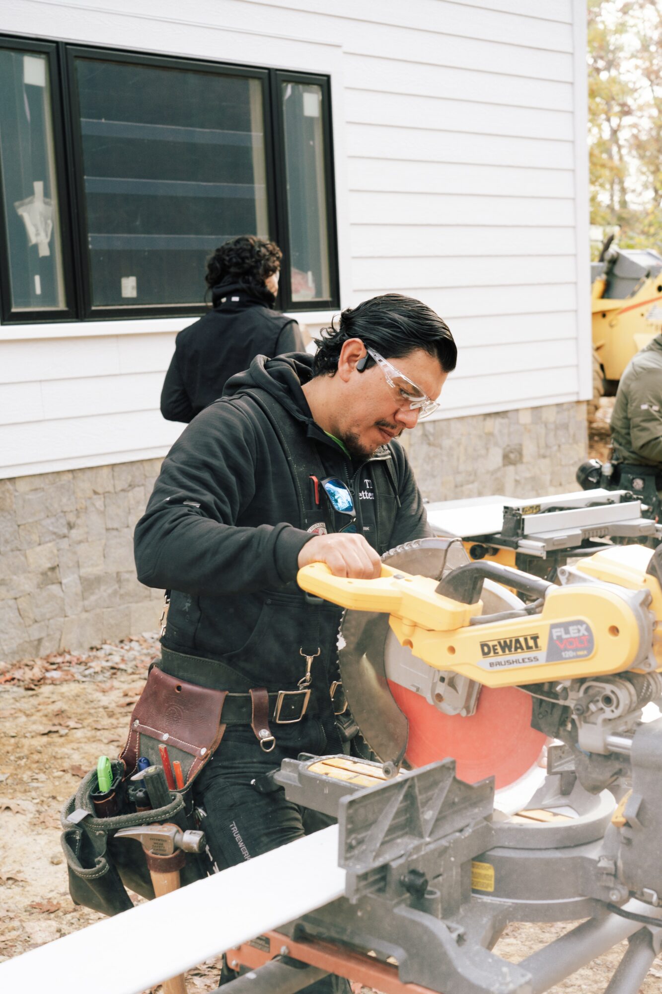 Man using a yellow DeWalt miter saw outdoors, wearing safety glasses and a black hoodie, with a white building in background.