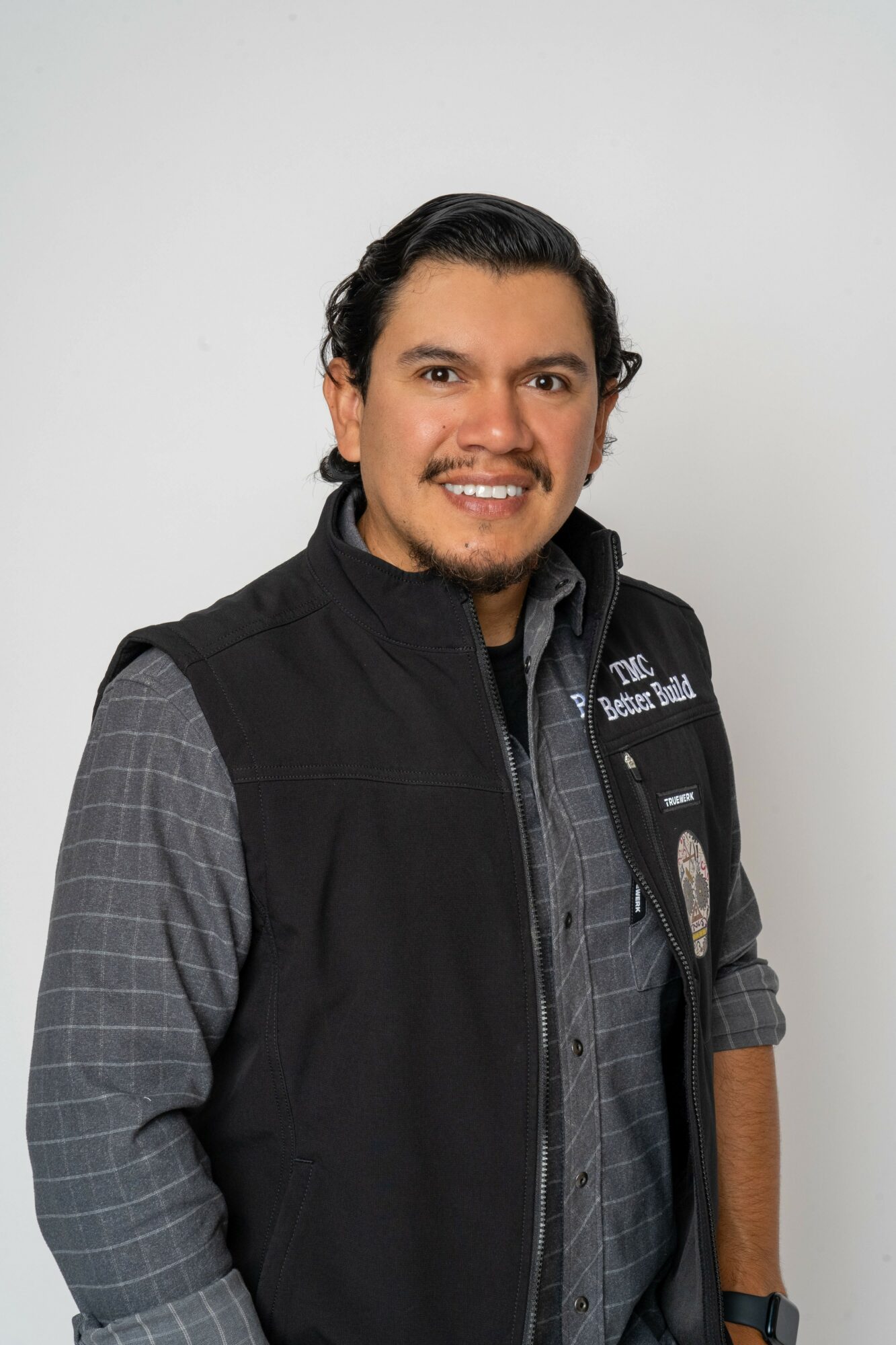 Man with dark hair and beard wearing a black vest over a gray checkered shirt, smiling.