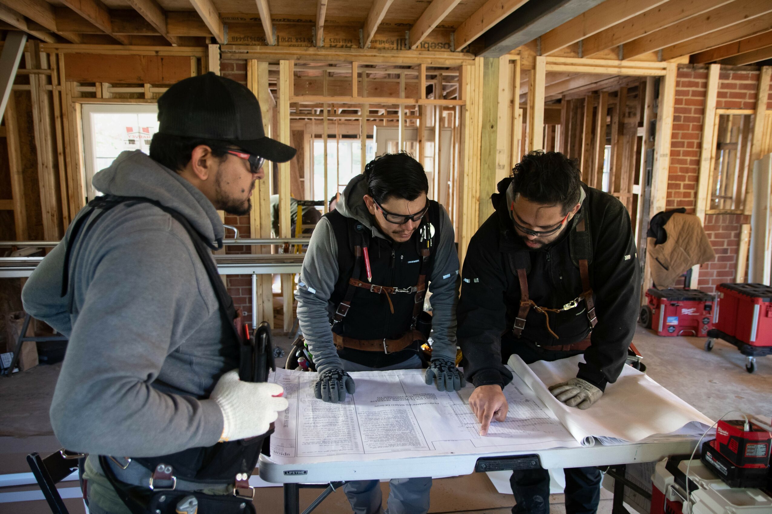 Three people examining blueprints on a construction site with wooden framing in the background.