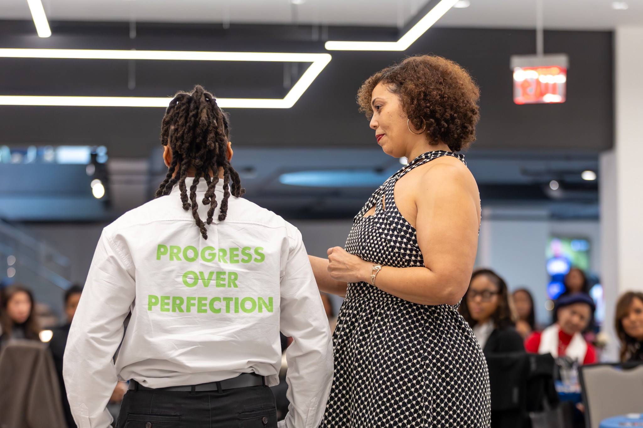 Two women engaged in conversation in a modern indoor setting with an audience in the background.