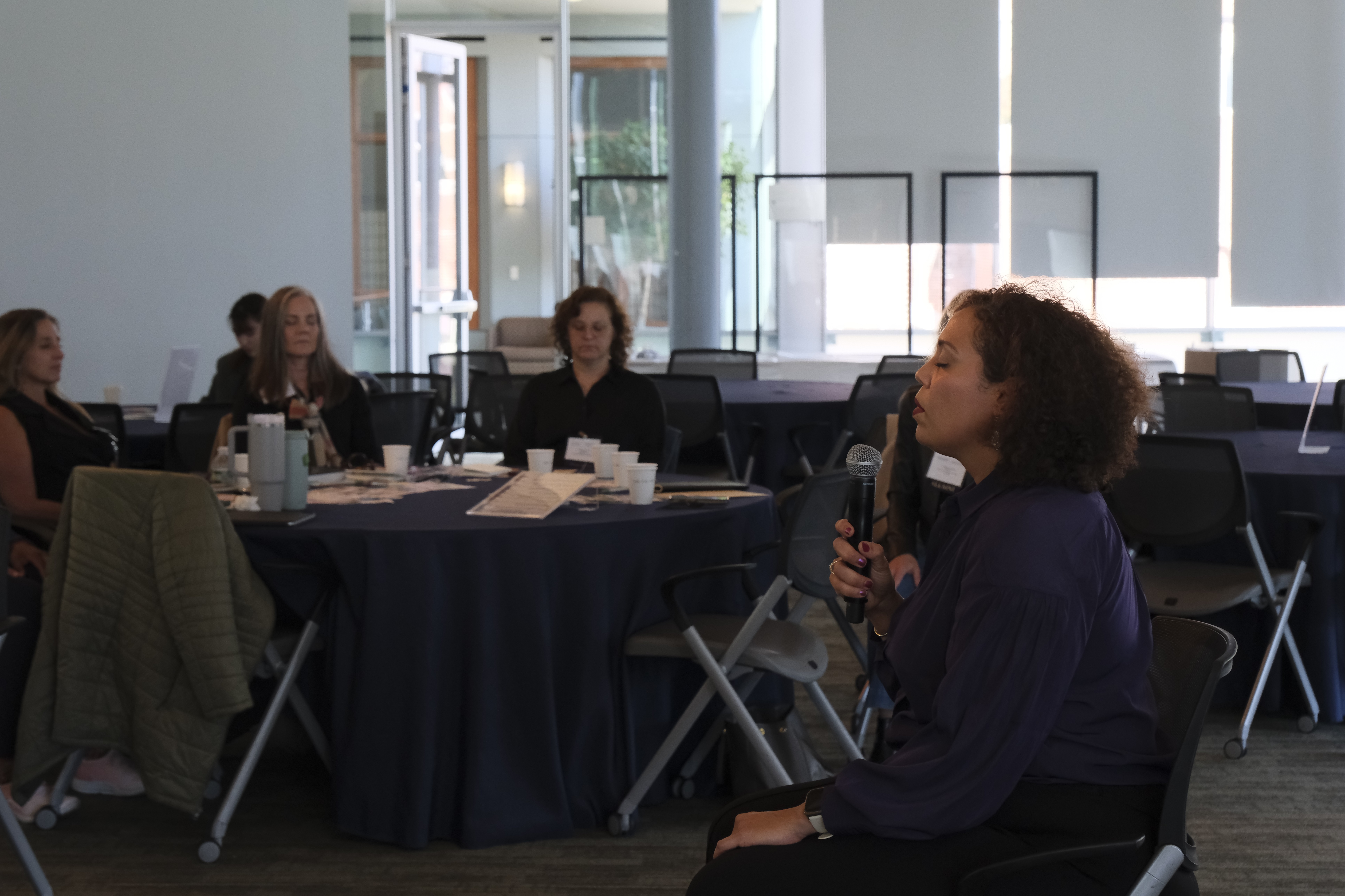 Woman speaking into microphone in a room with seated audience around a table.