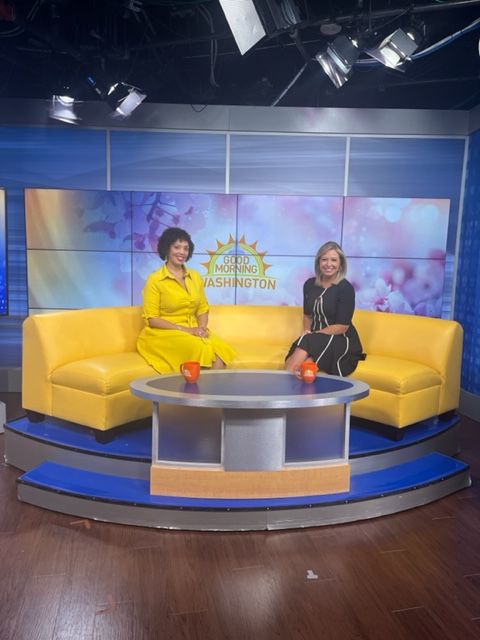 Two women sit on a yellow sofa in a TV studio with a large screen behind them displaying a logo and floral background.