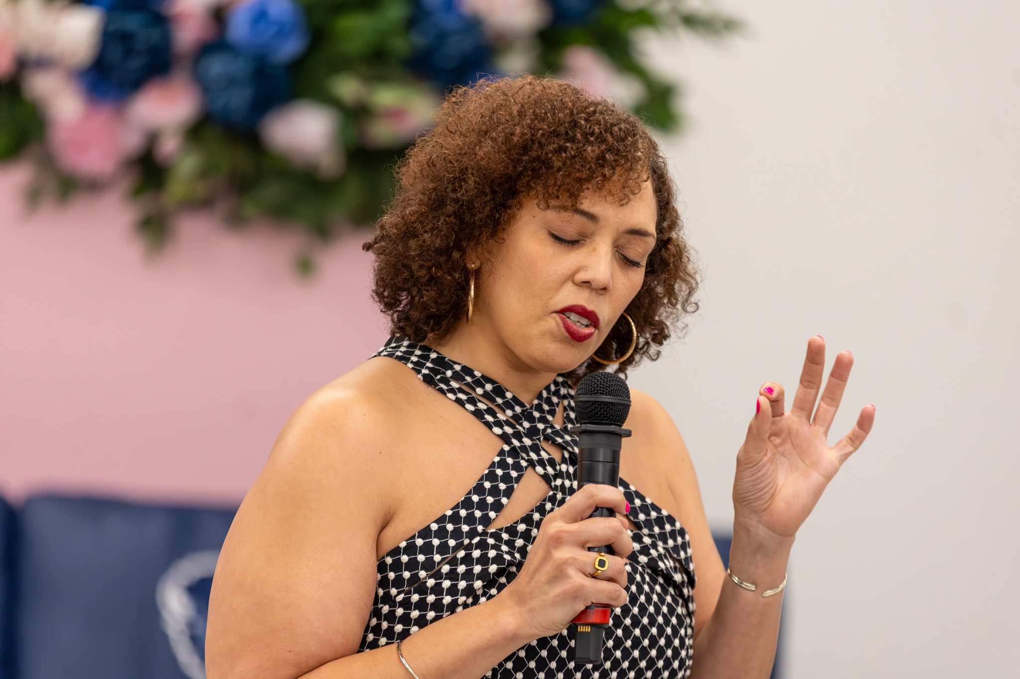 Woman with curly hair holding a microphone, eyes closed, in front of a floral background.