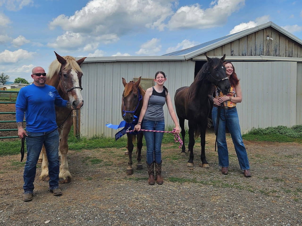 Two women and two men with three horses outdoors near a building, some holding horse leads, on a partly cloudy day.