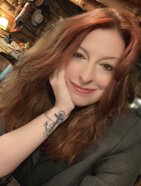 Woman with long, wavy red hair smiling, resting her face on her hand, in a cozy indoor setting.