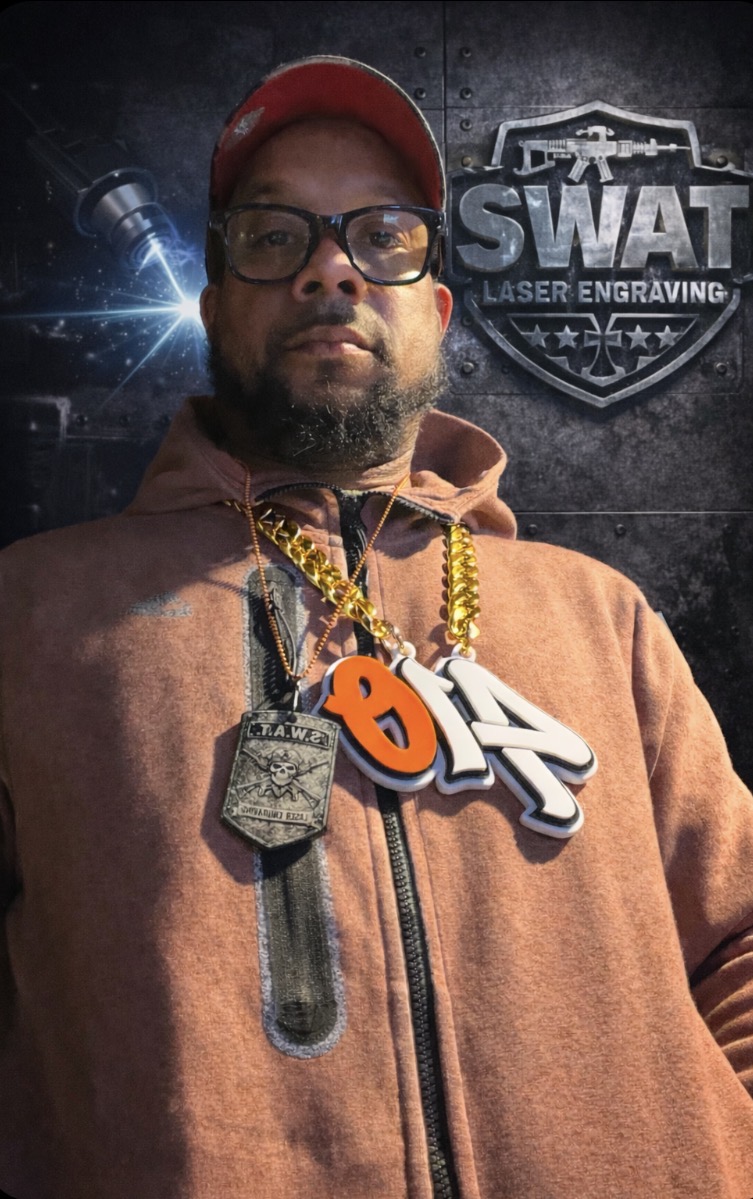 Man wearing glasses, red cap, orange hoodie, and large chain with a badge and orange and white pendant, standing in front of SWAT sign.