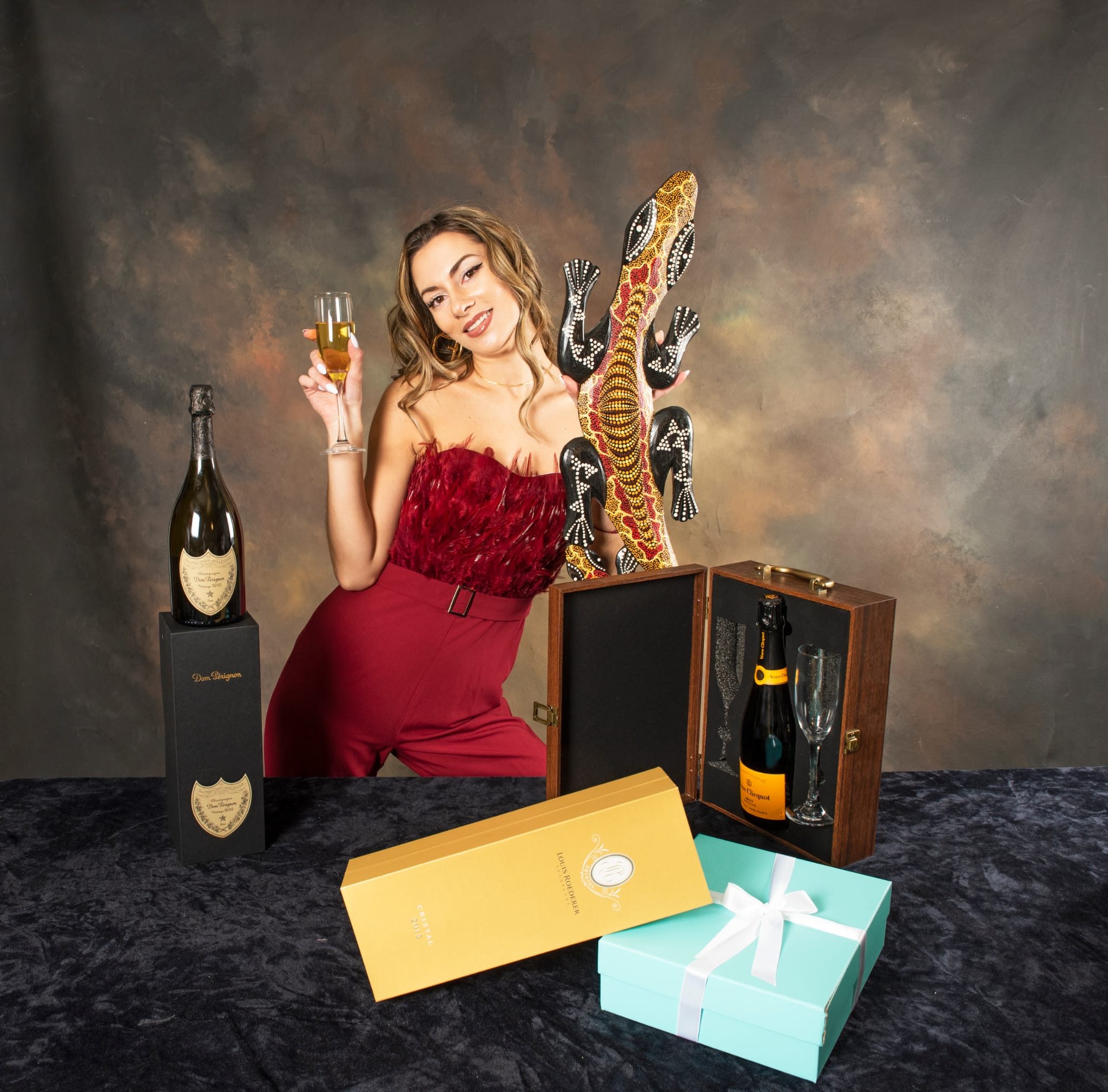 Woman in red dress holding a glass, surrounded by champagne bottles and gift boxes, smiling at camera.