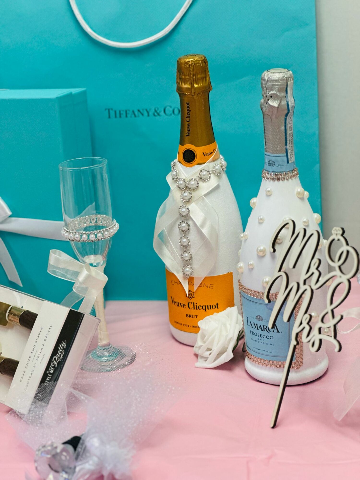 Two decorated champagne bottles with pearl necklaces, a glass of champagne, and a wedding sign on pink fabric.