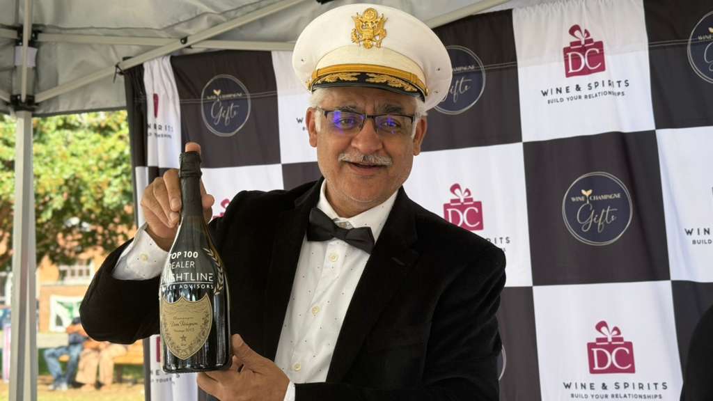 Man in a white captain's hat and black tuxedo holding a large bottle of wine or champagne.