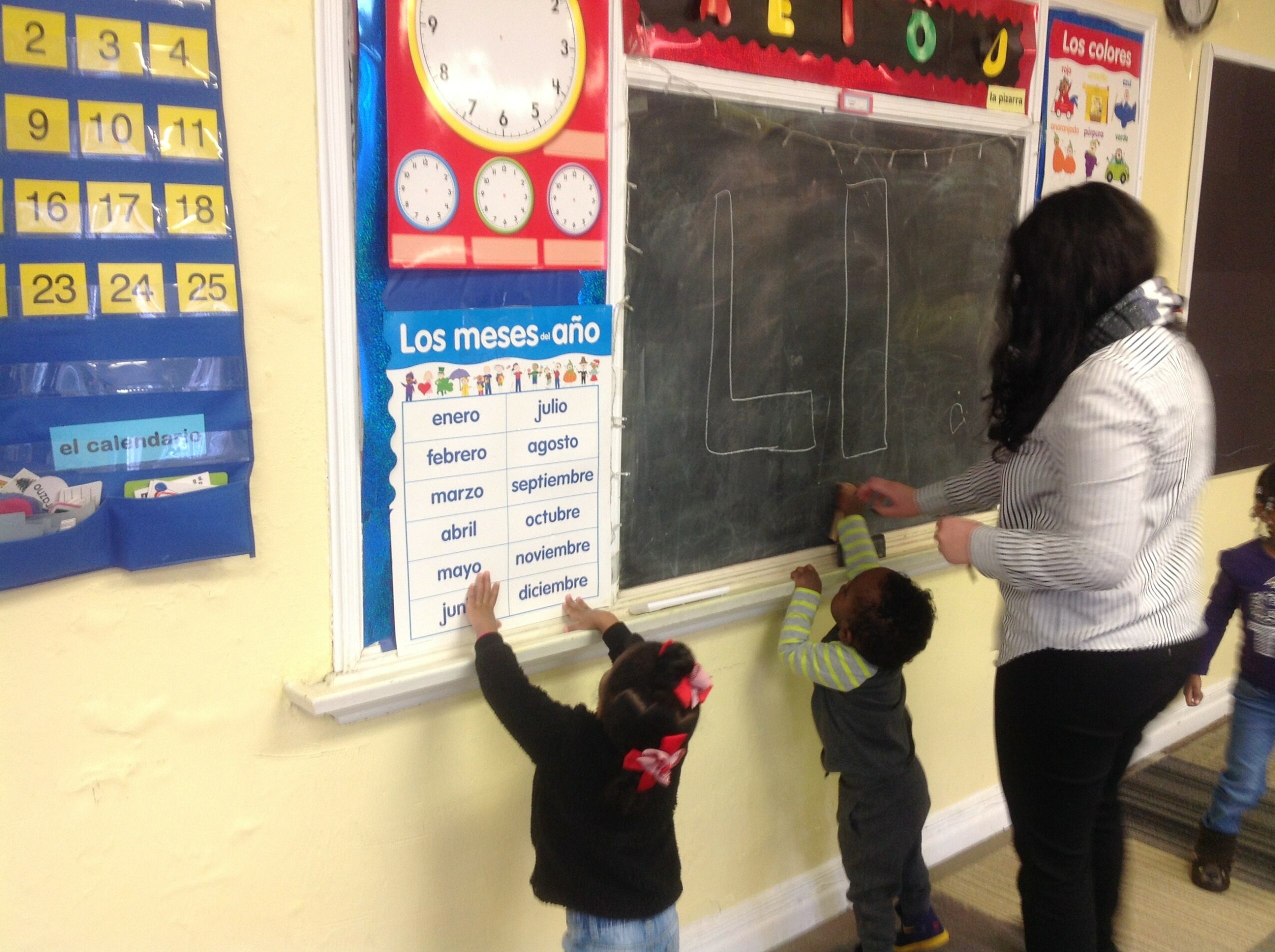 Teacher and children at blackboard, writing and drawing, classroom setting with educational posters and calendar.