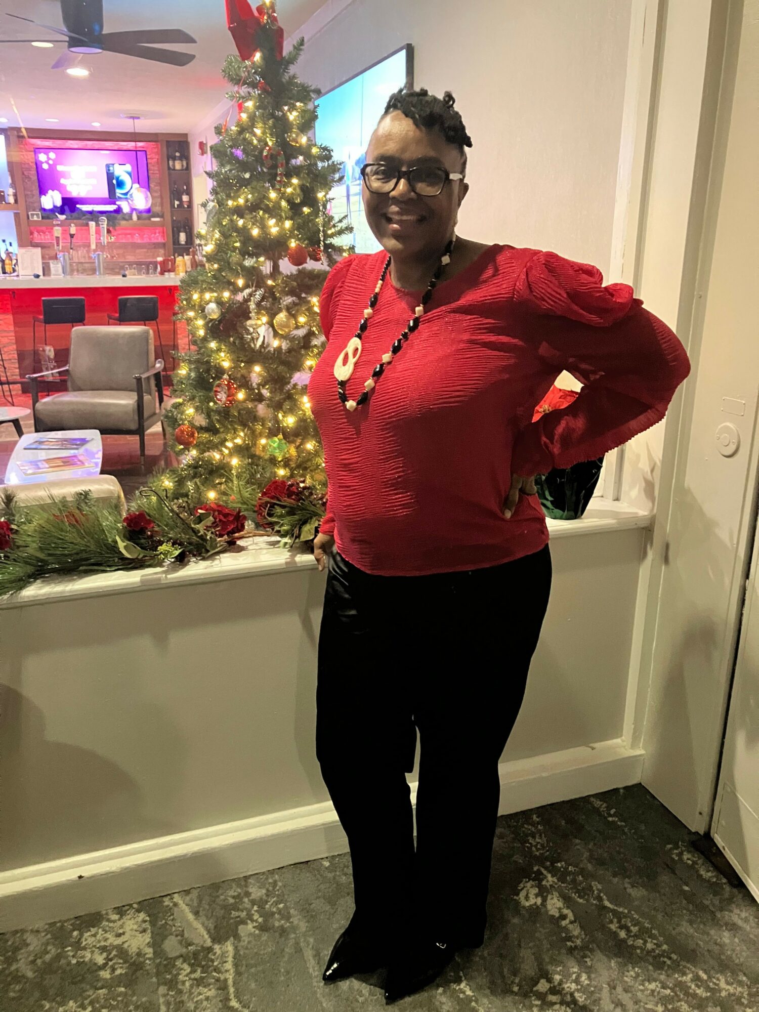 Woman in red top and black pants standing indoors near decorated Christmas tree with lights and ornaments.