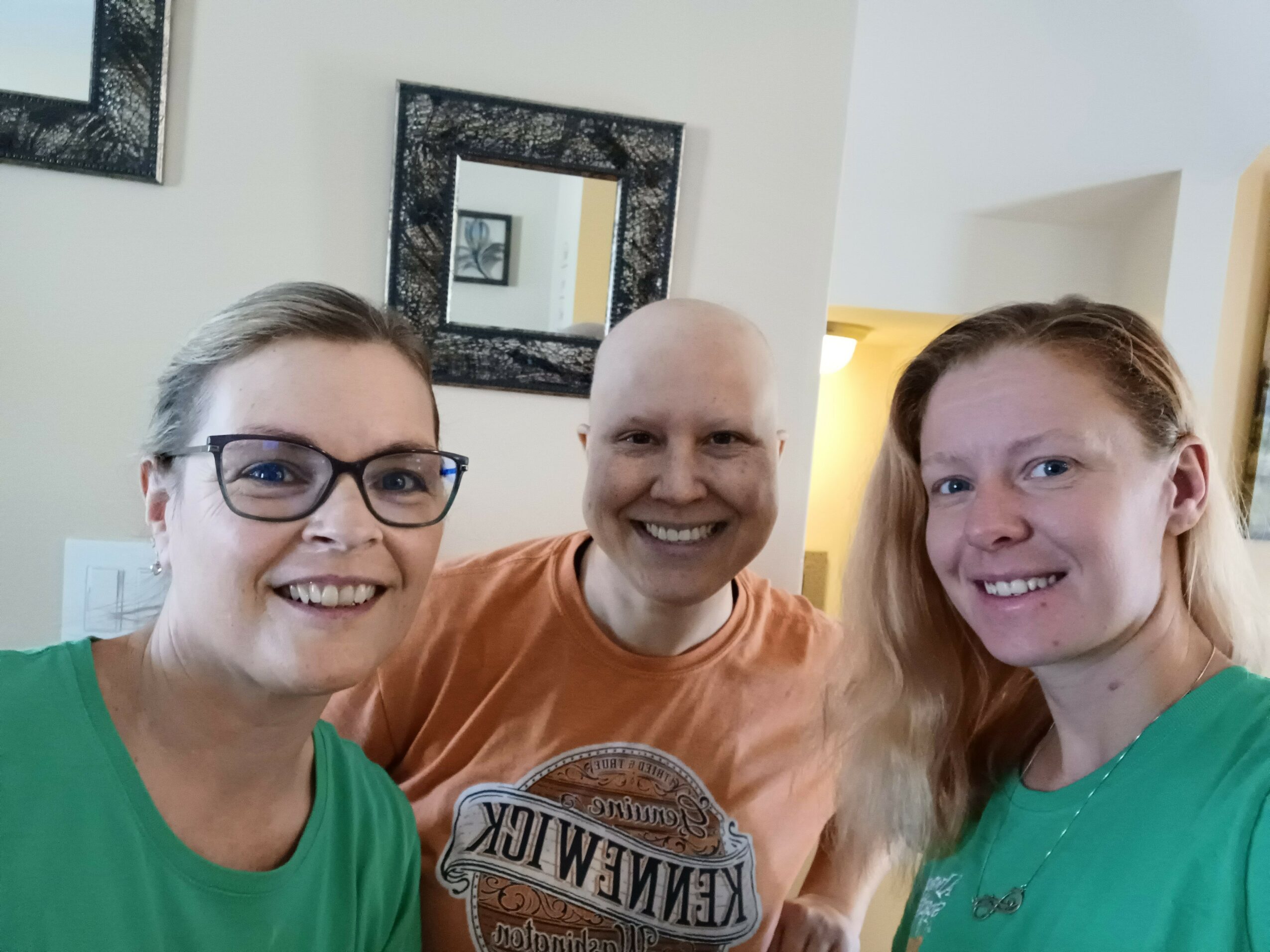 Three smiling women taking a selfie indoors, two wearing green shirts, one in orange, with framed pictures on the wall behind.