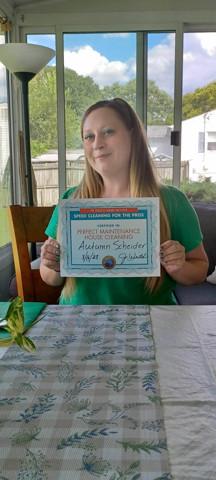 Woman holding a certificate at a table with a window view of trees and houses behind her.
