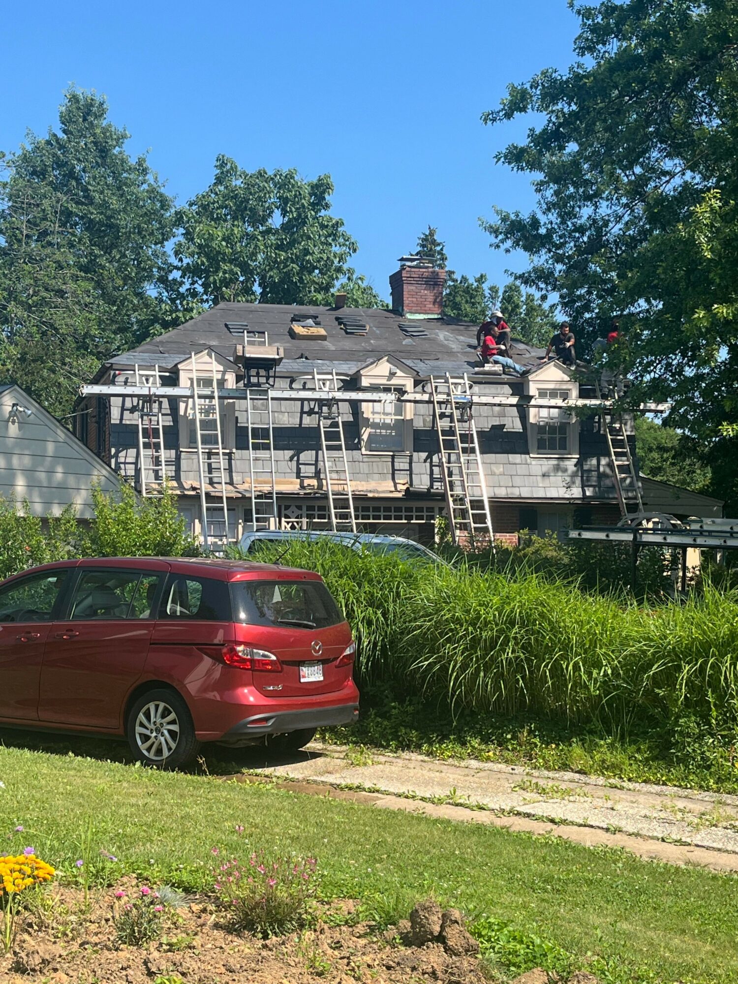 House under renovation with scaffolding, trees in background, red car parked on grass, blue sky above.