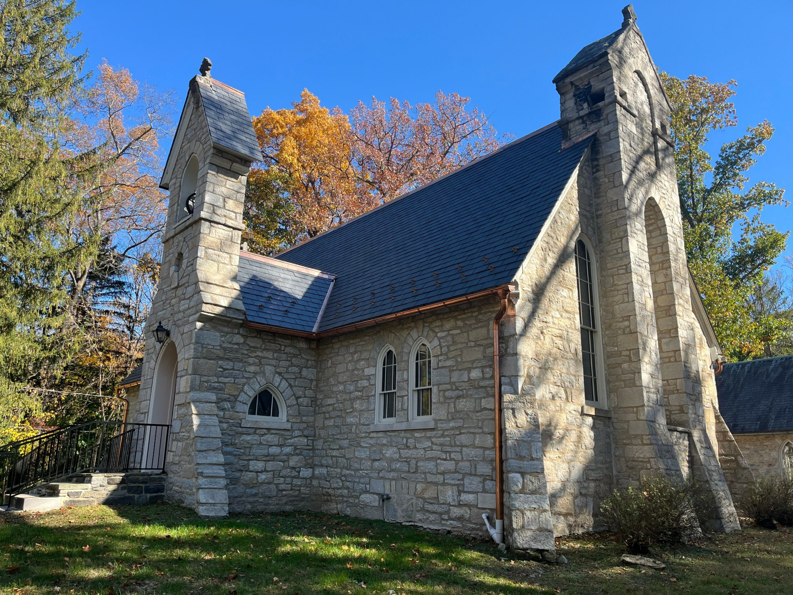 Stone church with pointed towers, arched windows, surrounded by trees with autumn foliage, under a clear blue sky.