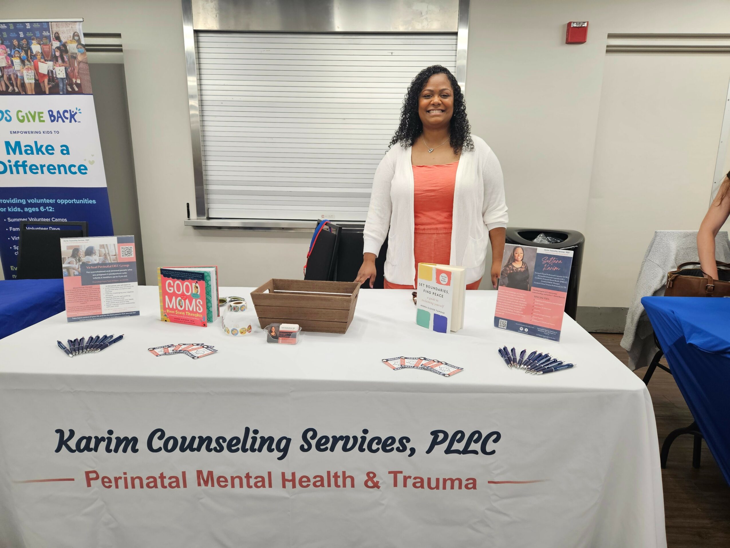 Woman standing behind table with promotional materials, in room with closed blinds, promoting counseling services.
